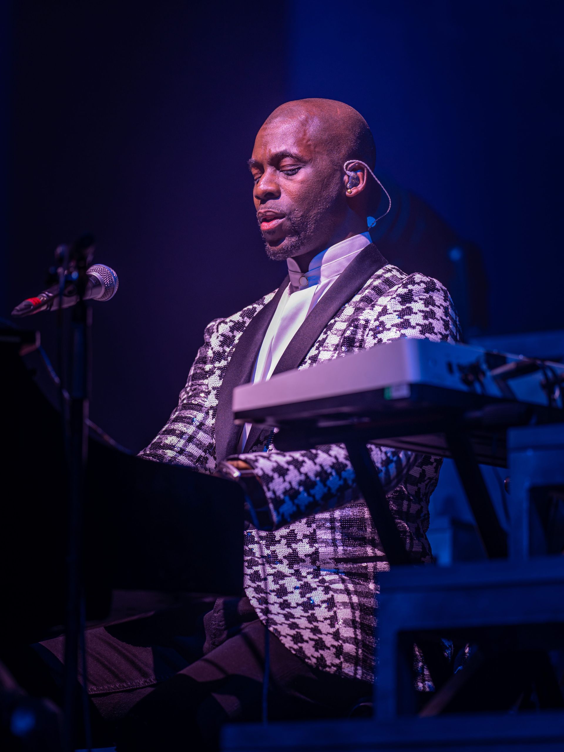 Keyboardist on stage wearing a black and white houndstooth jacket and white shirt. Dark setting with blue lighting.