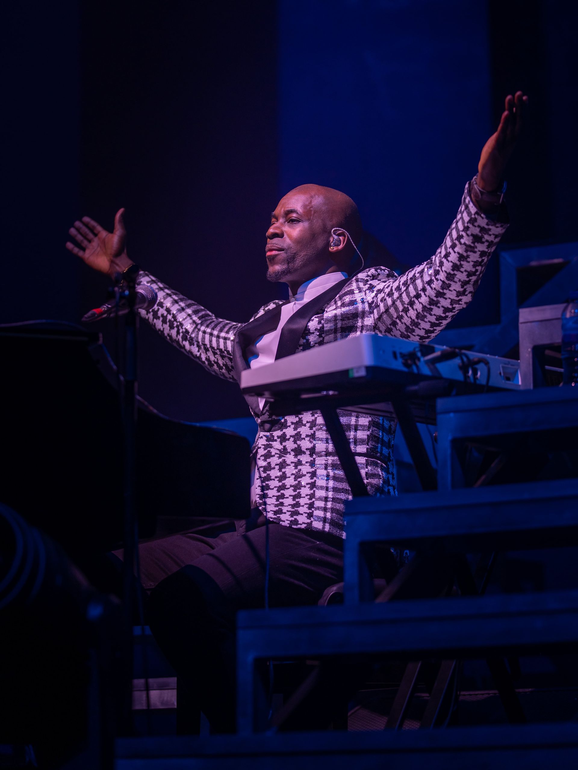 Keyboardist in black and white houndstooth jacket with arms raised, lit by blue stage lights.