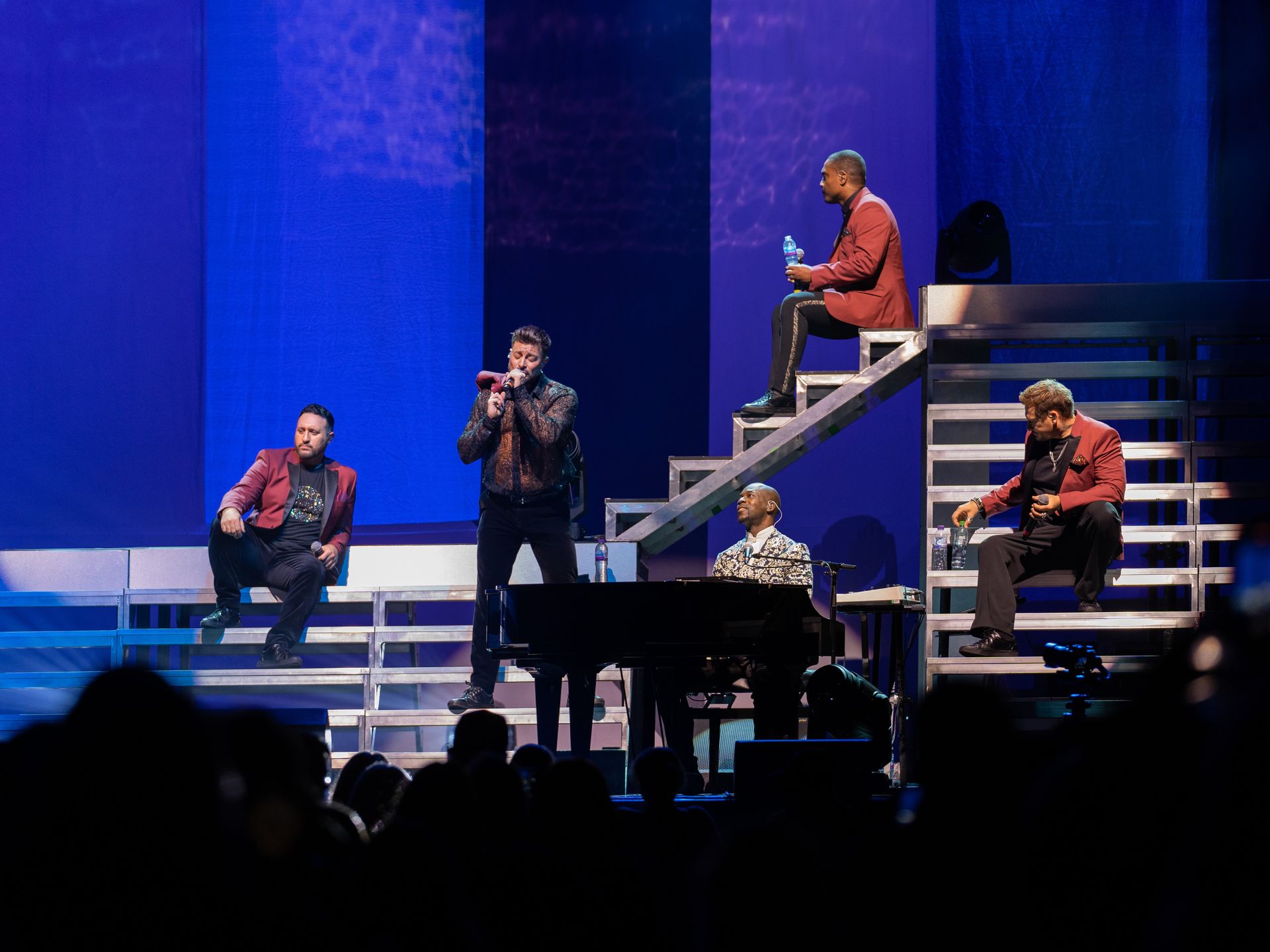 Boy band performing on stage with blue backdrop, some members on a staircase.