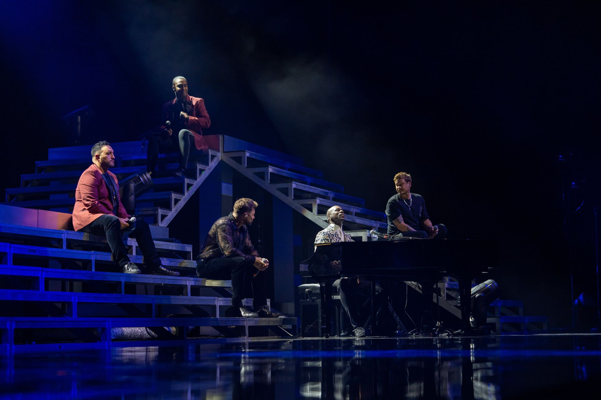 Five men on a stage with stairs; one plays keyboard, others sit or stand. Blue lighting.