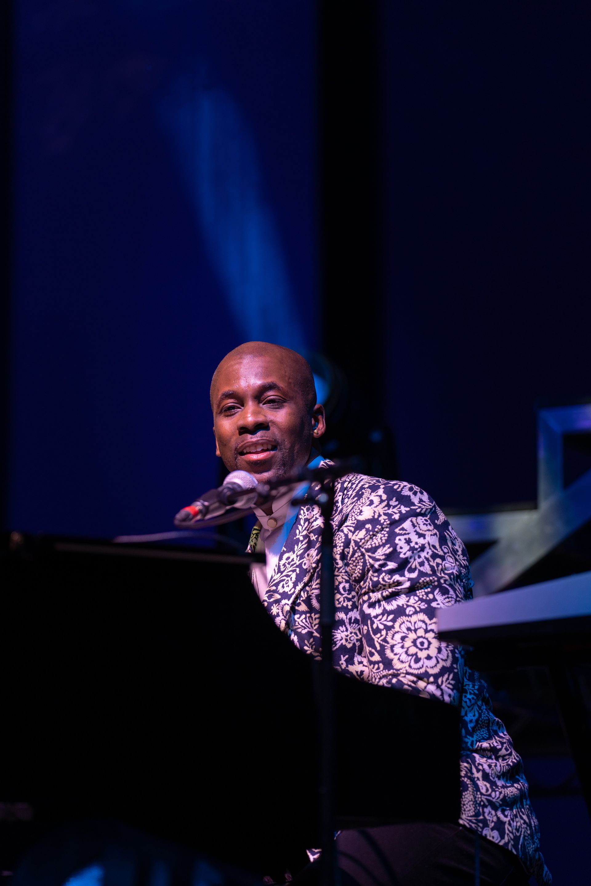 Man playing piano on stage, wearing patterned jacket. Blue stage lighting.