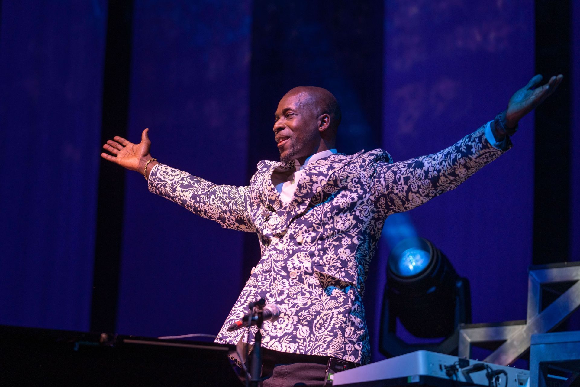 Man on stage with arms outstretched, wearing a patterned jacket, lit by blue stage lights.