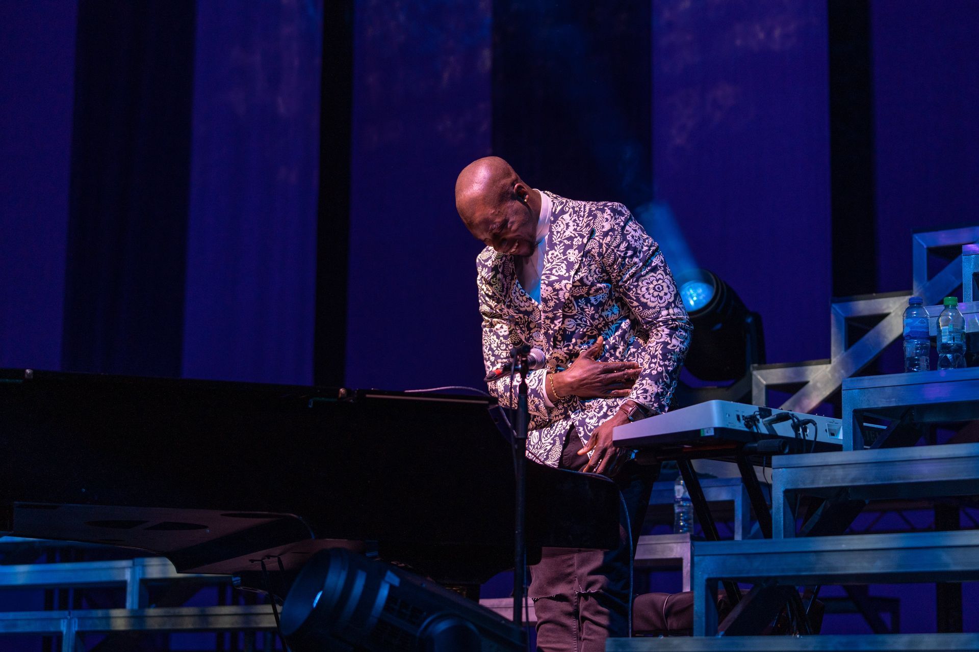 A man in a patterned shirt plays a keyboard on stage, illuminated by blue lighting.