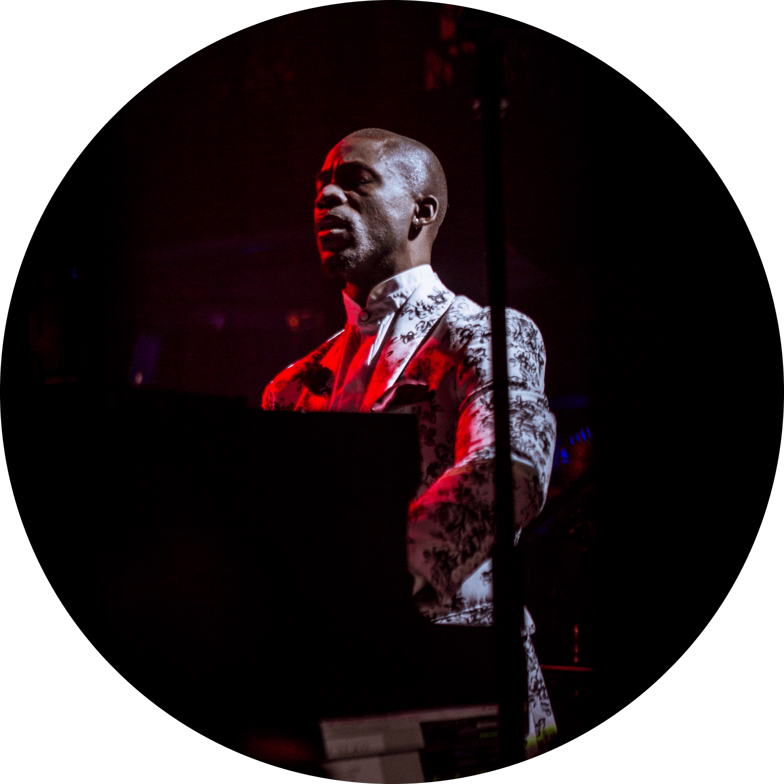 A man in a white patterned suit performing on a keyboard under red stage lights.