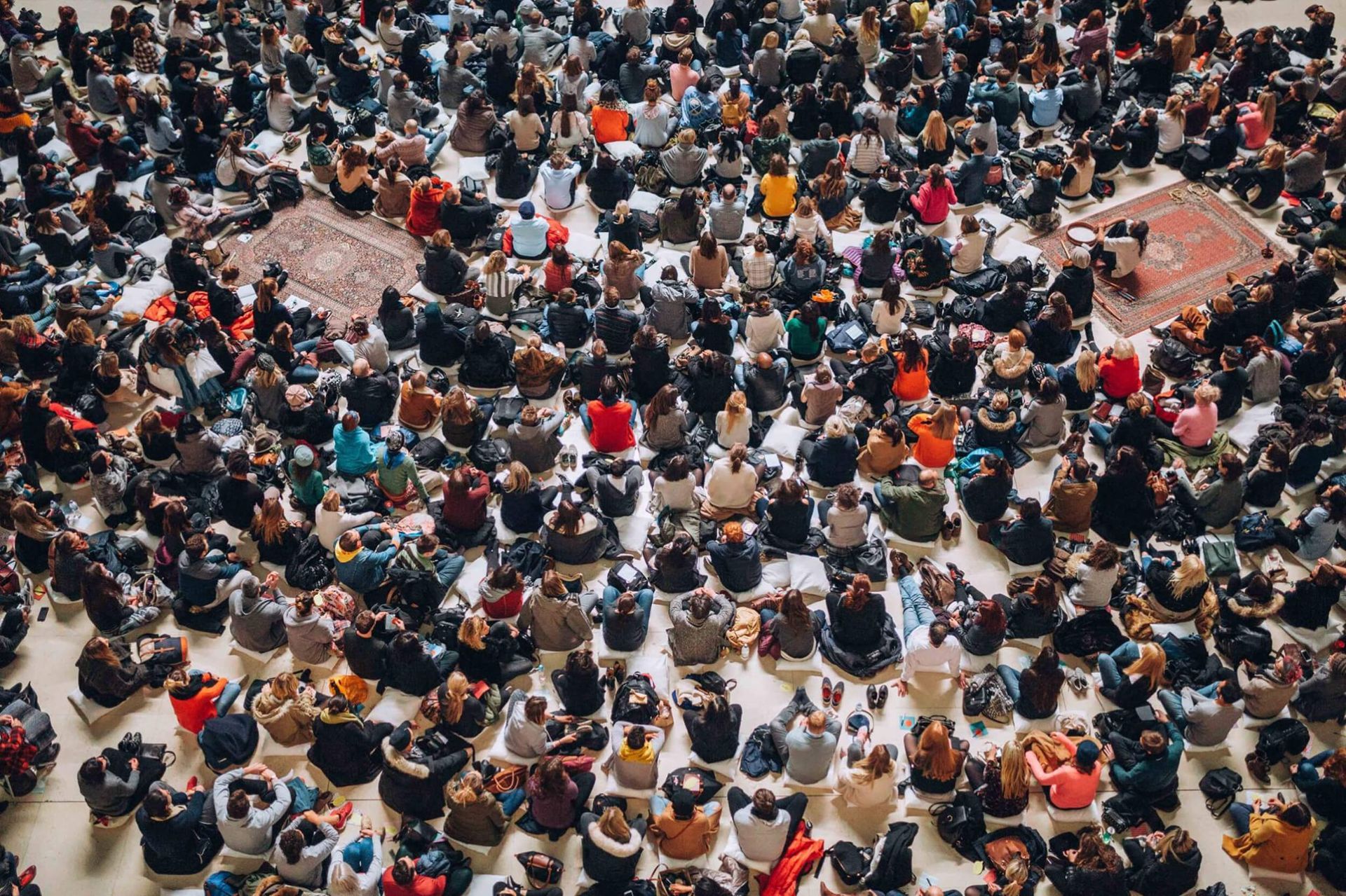 Overhead view of a large crowd of people seated on the floor, some on rugs.