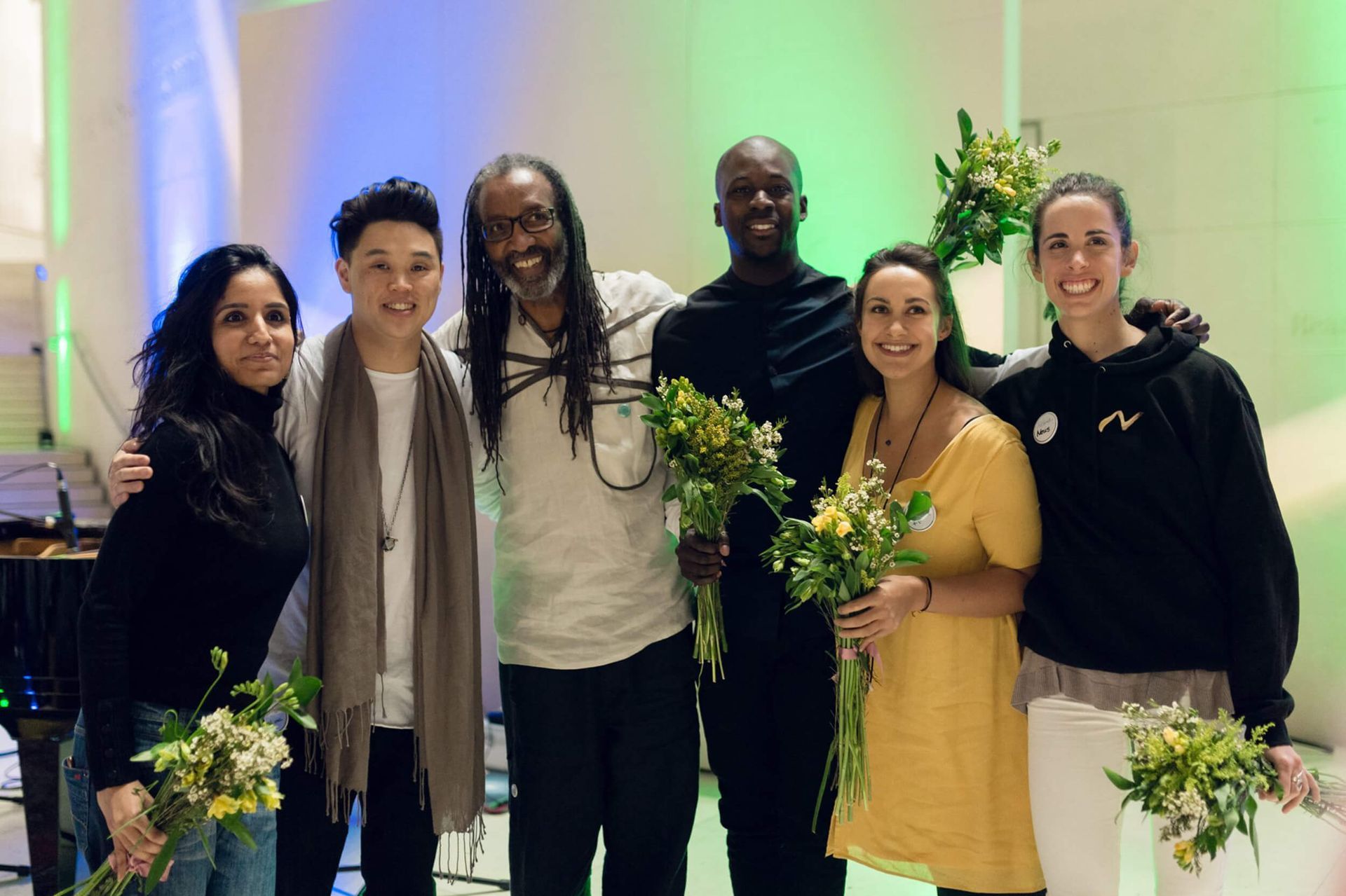 Group of six people smiling, holding bouquets, with green and blue lighting.