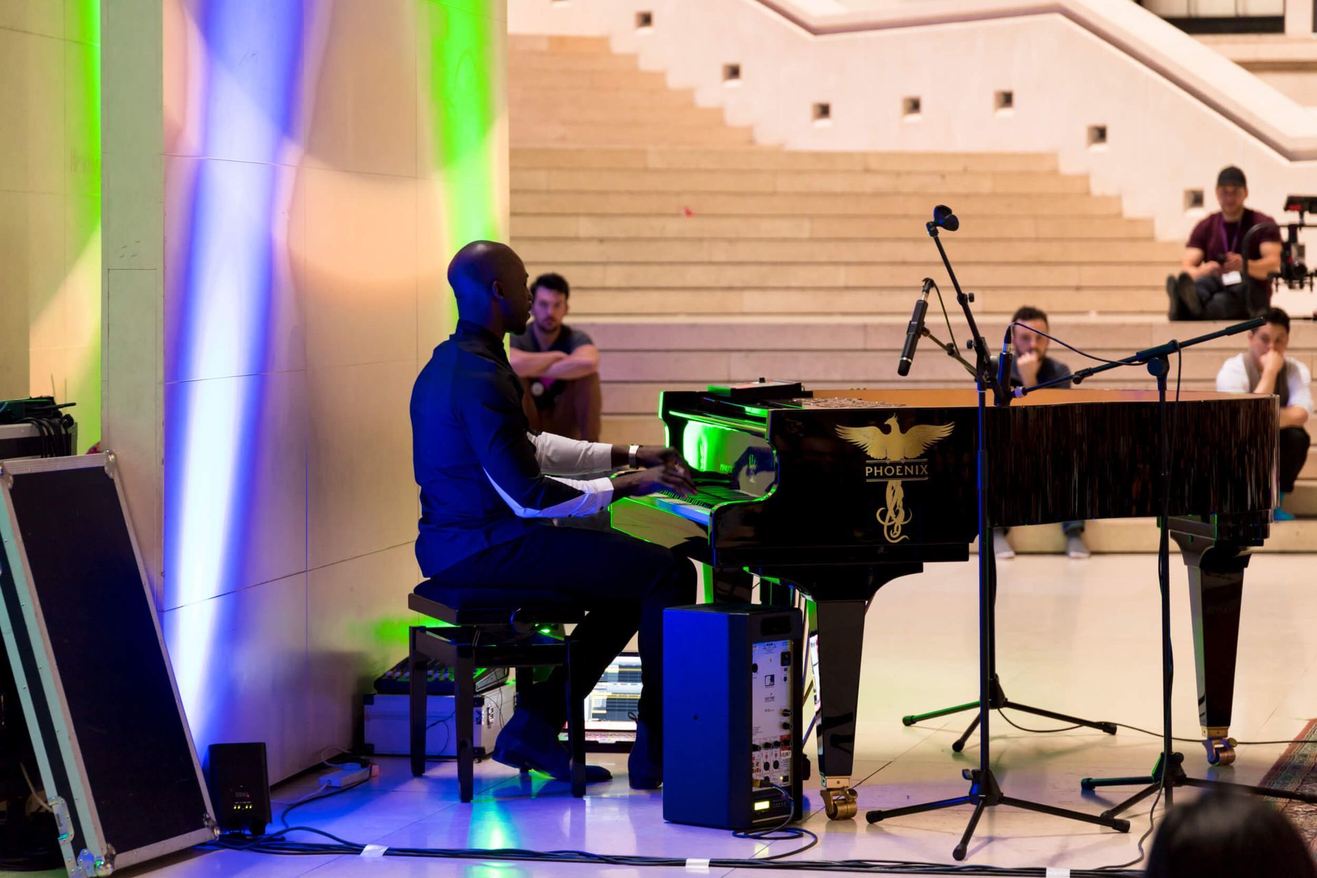 Pianist at keyboard performs onstage with microphones, audience, and colorful stage lighting near stairs.