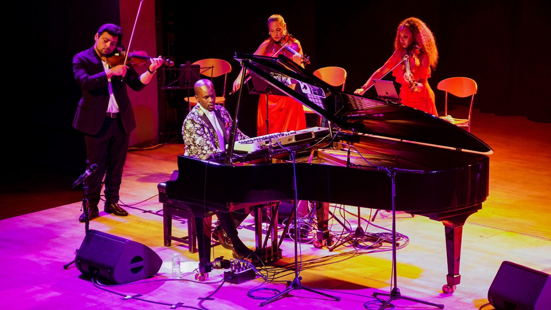 A musical ensemble performs onstage: piano, violins, and a violinist playing with animated energy under stage lights.