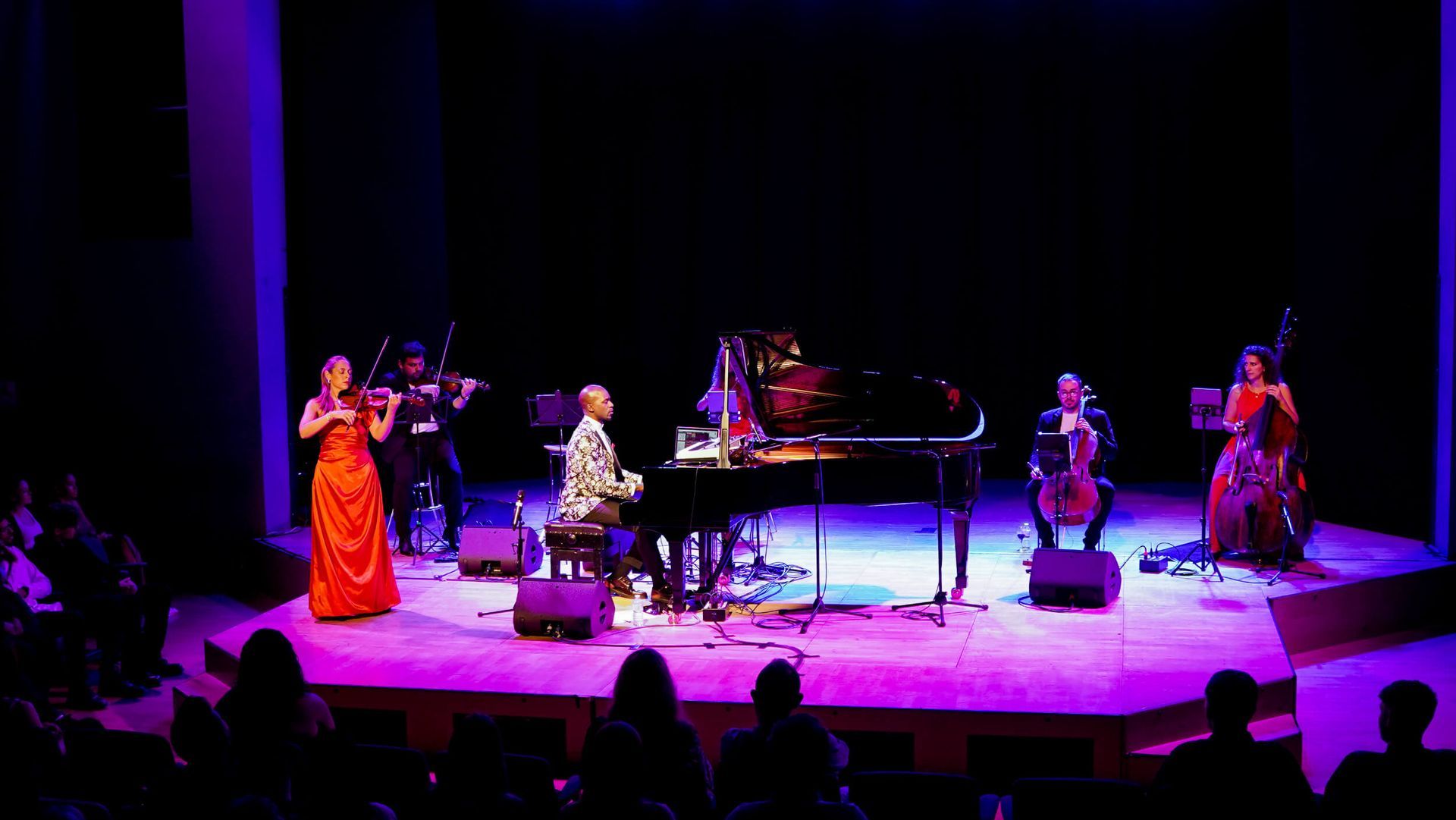 Musicians on stage: piano, violin, cello, bass. Purple lighting. Audience in foreground.