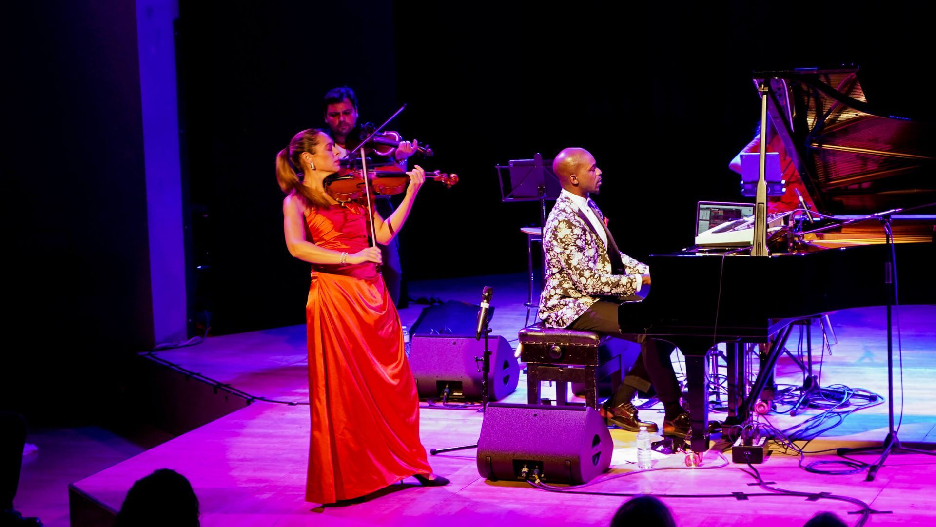 Musicians perform on stage: violinist in red dress, pianist in the background. Stage lit with blue and purple light.