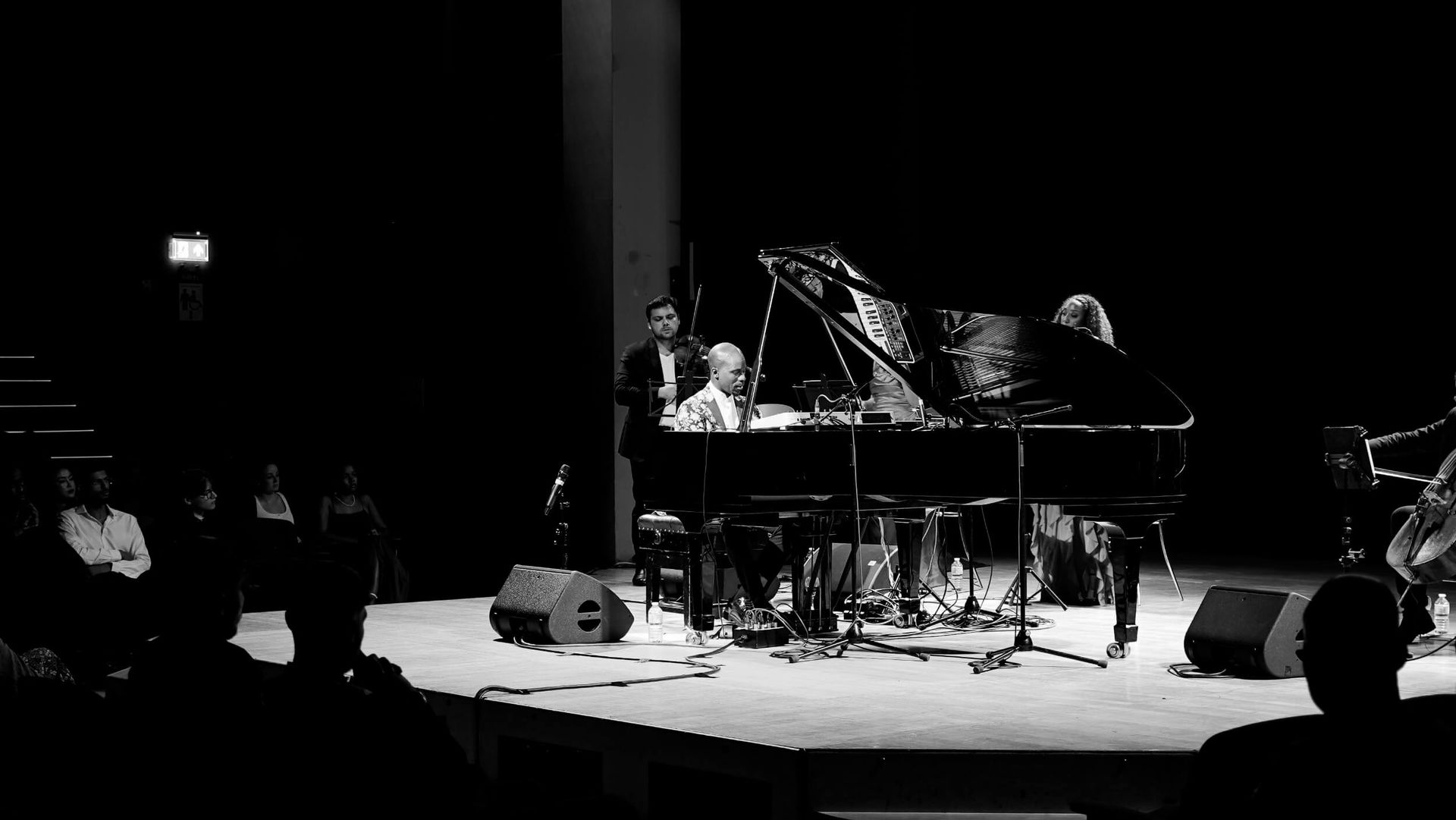 A jazz performance. Musicians on stage with a grand piano. Audience members in the foreground. Black and white.