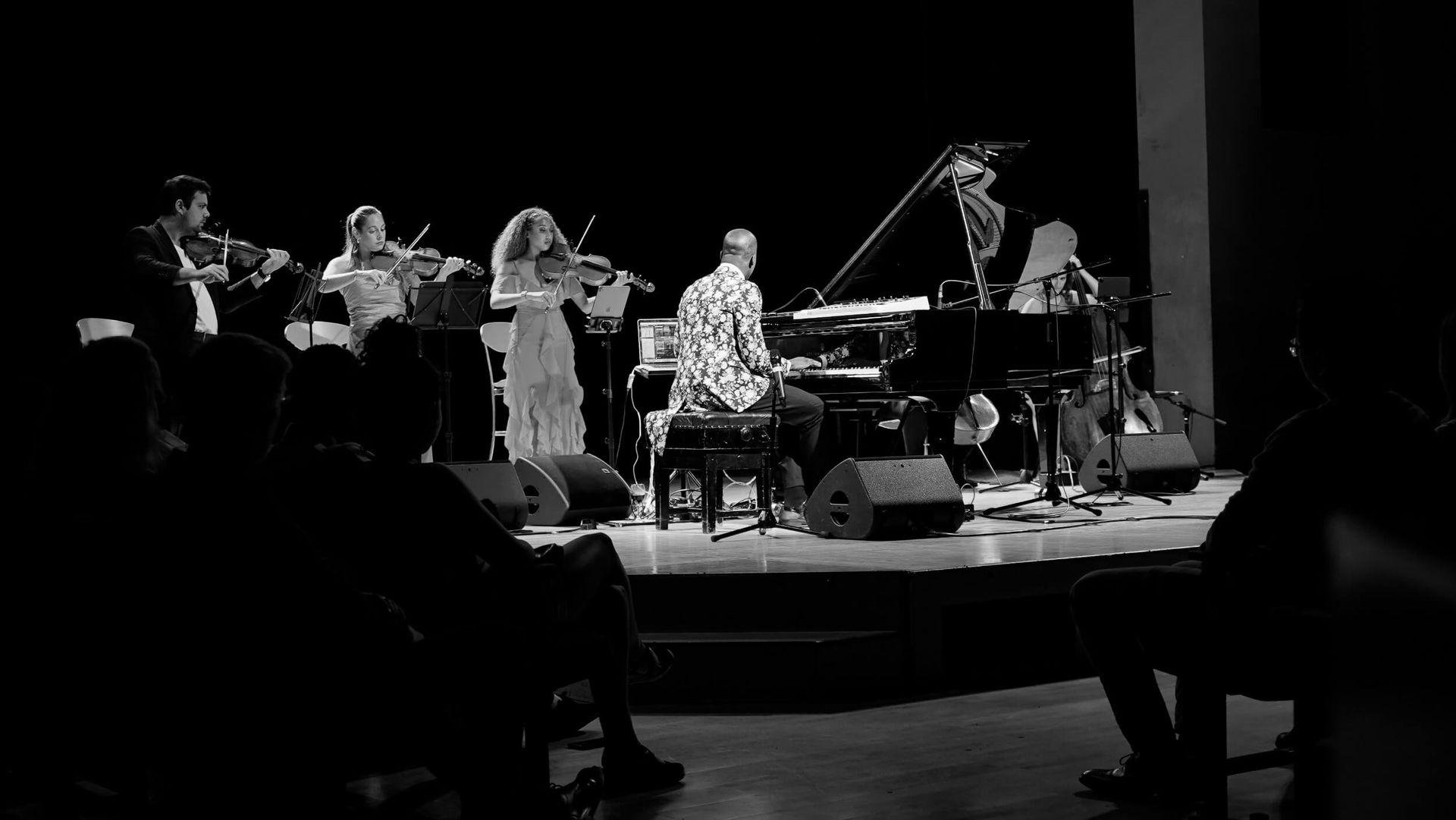 Musical ensemble on stage, playing instruments. Pianist center, violinists, and audience silhouettes.