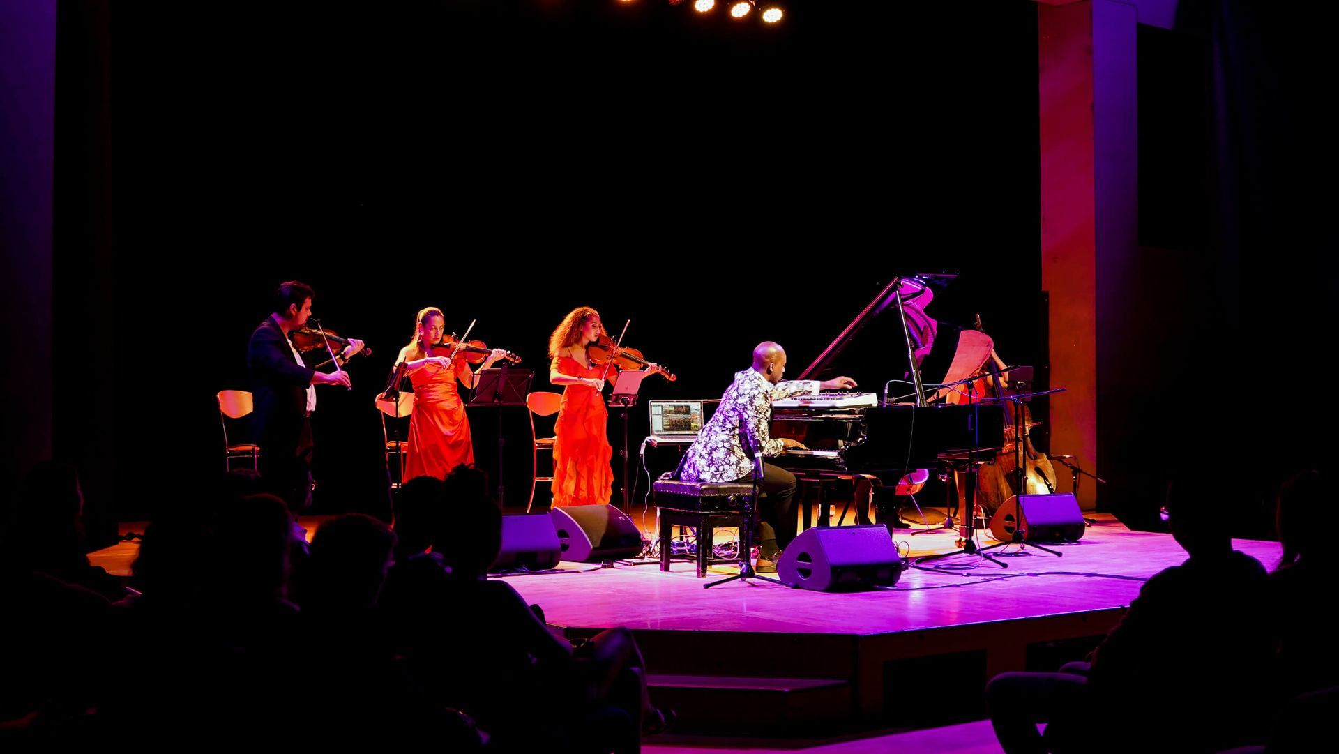 Musical ensemble on stage performing; strings and piano. Red and purple lighting. Audience visible.