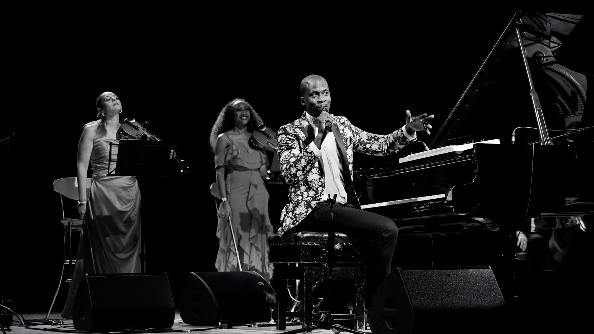 A musician sings at a piano, with a violinist and vocalist on stage. Black and white.
