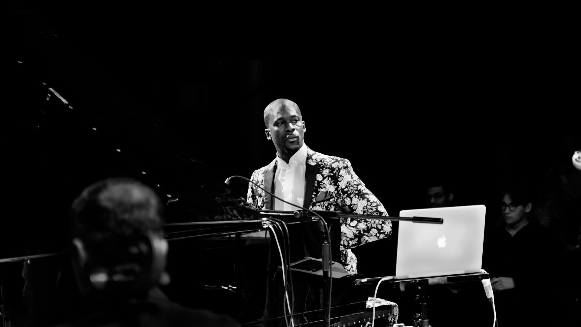 Man in patterned blazer at a piano, laptop nearby, on stage.