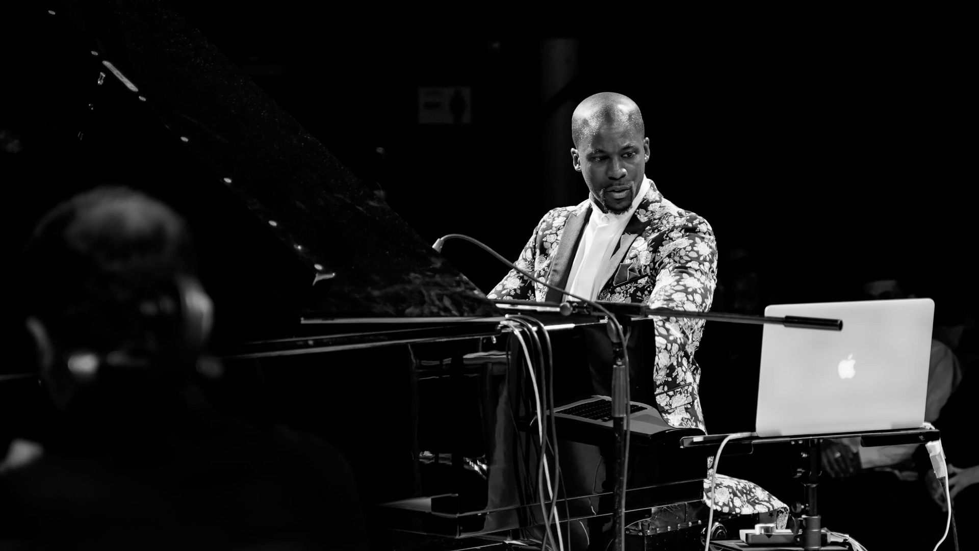Pianist in floral jacket performing at a concert, laptop on stand to right. Black and white.