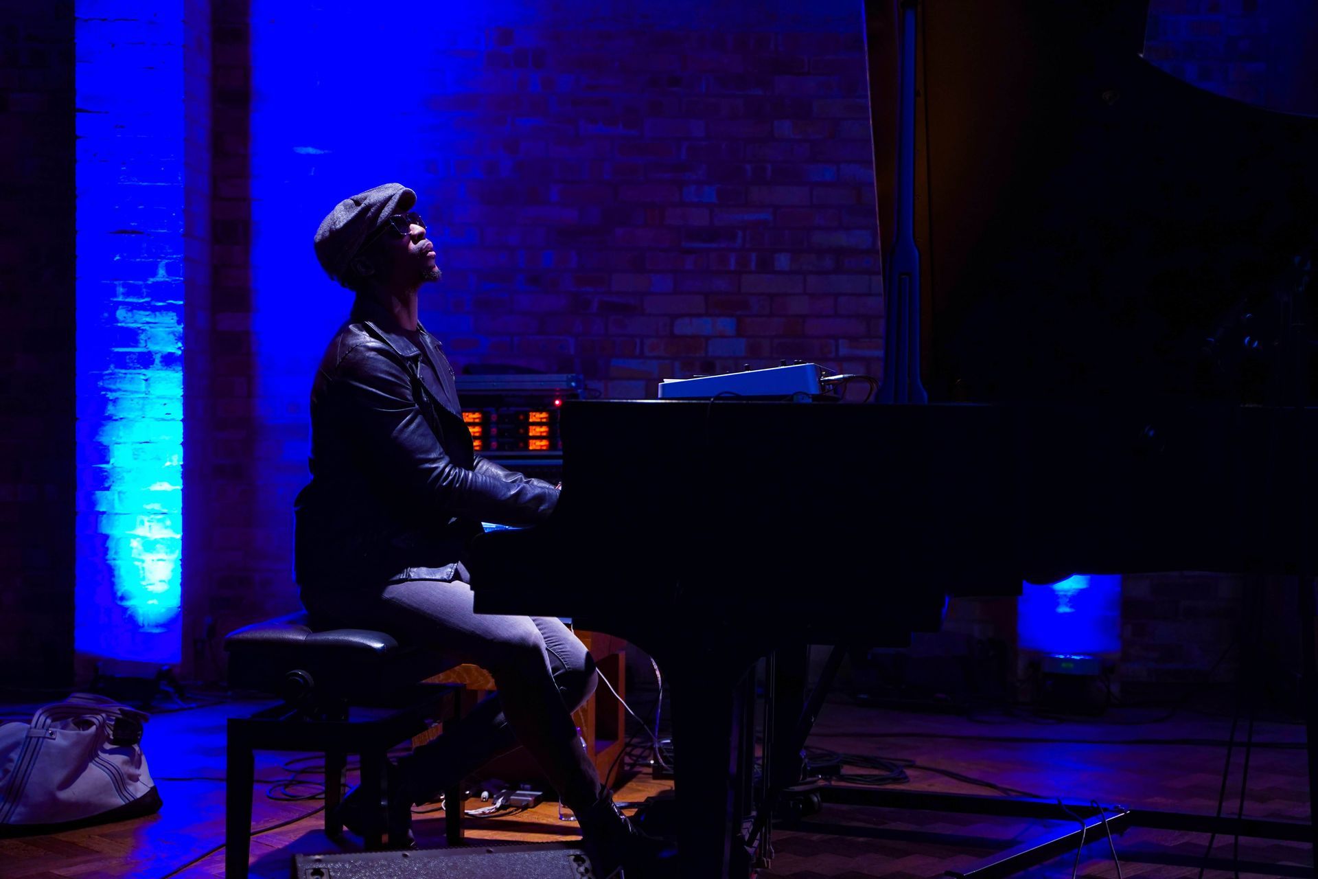 Person playing piano, lit with blue stage lights. Brick wall background.