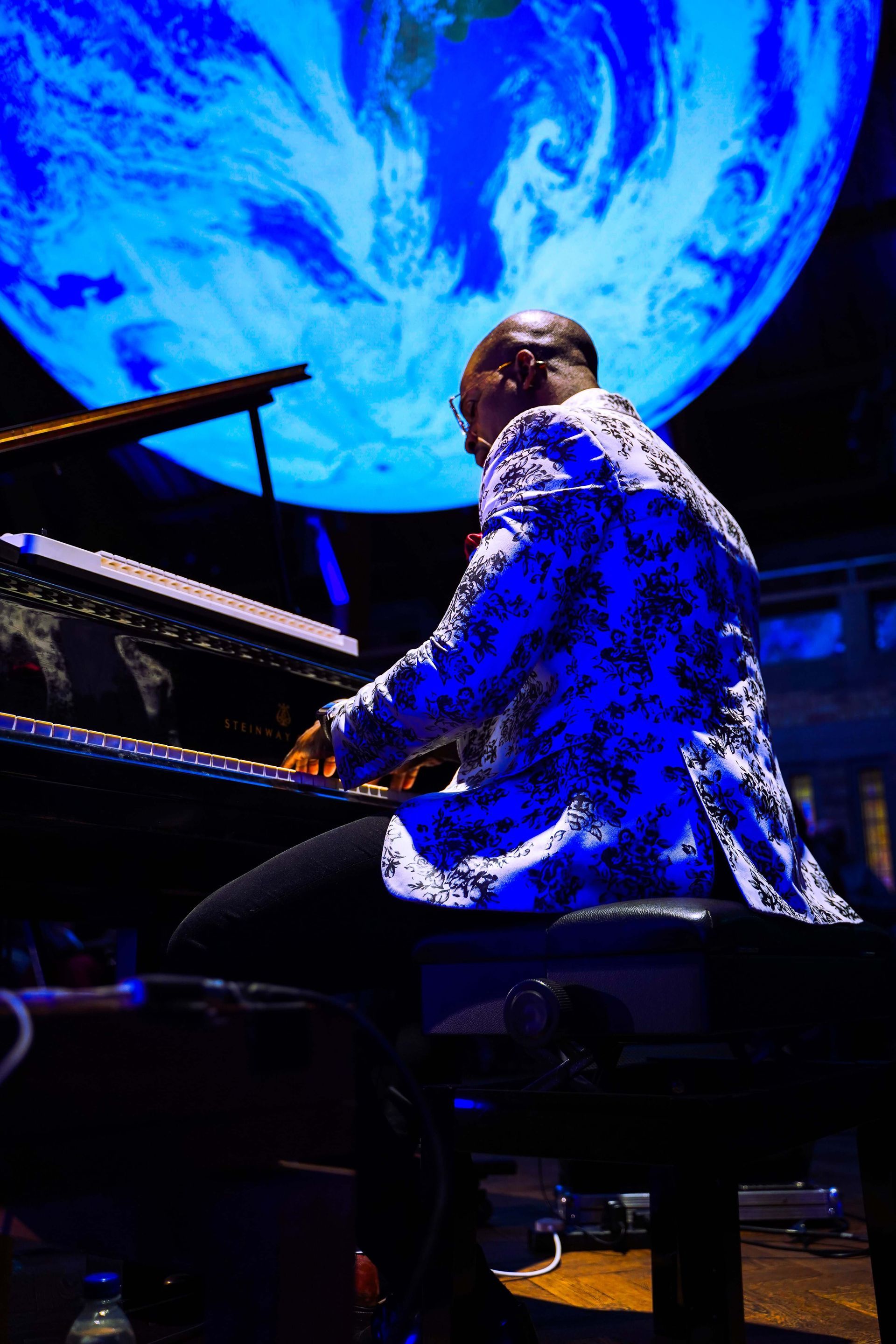 Pianist in a blue patterned jacket plays a grand piano, against the backdrop of a blue Earth projection.