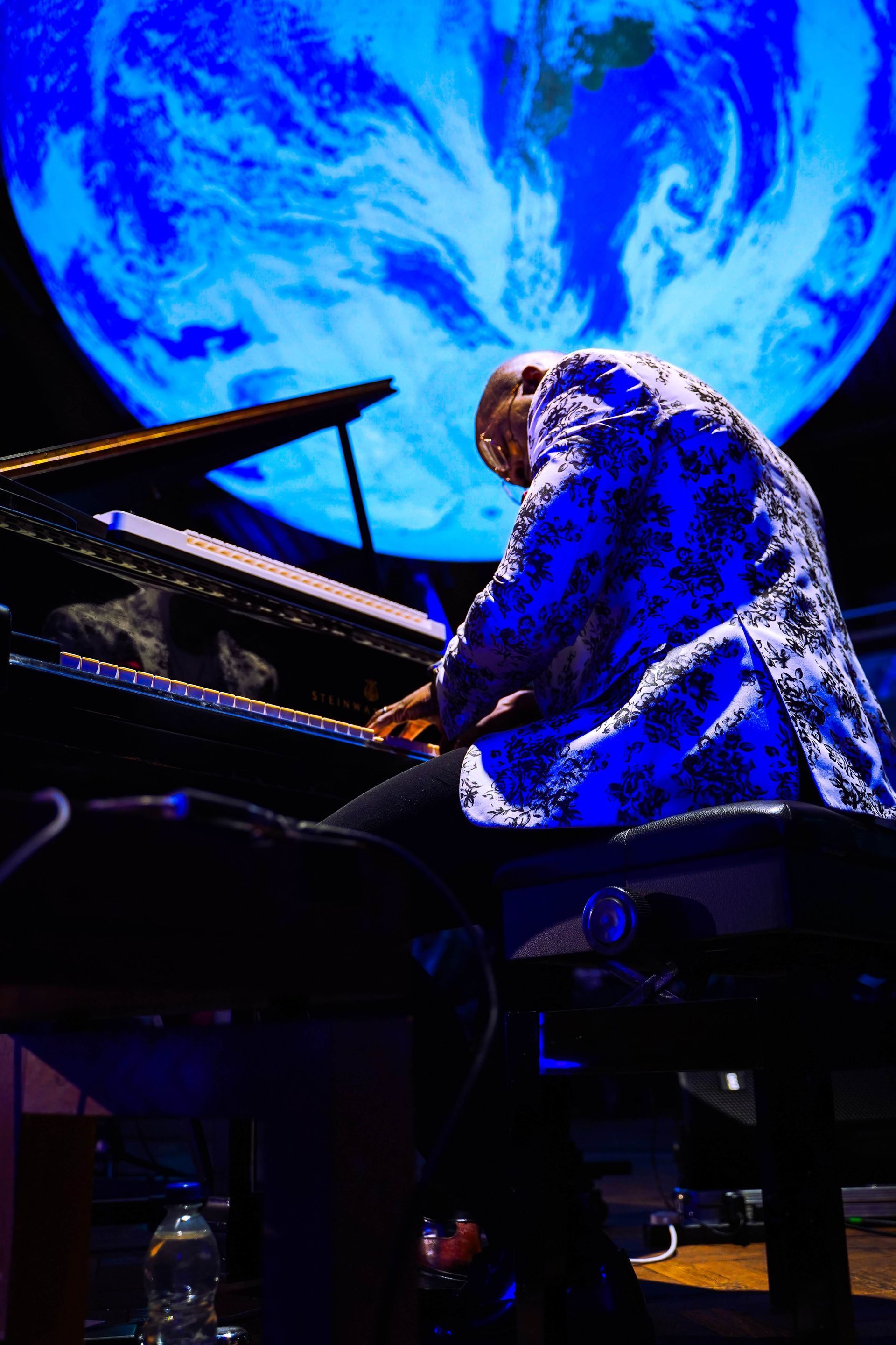 Pianist playing a grand piano. Blue-lit Earth in the background. Man in patterned jacket. Dark stage.