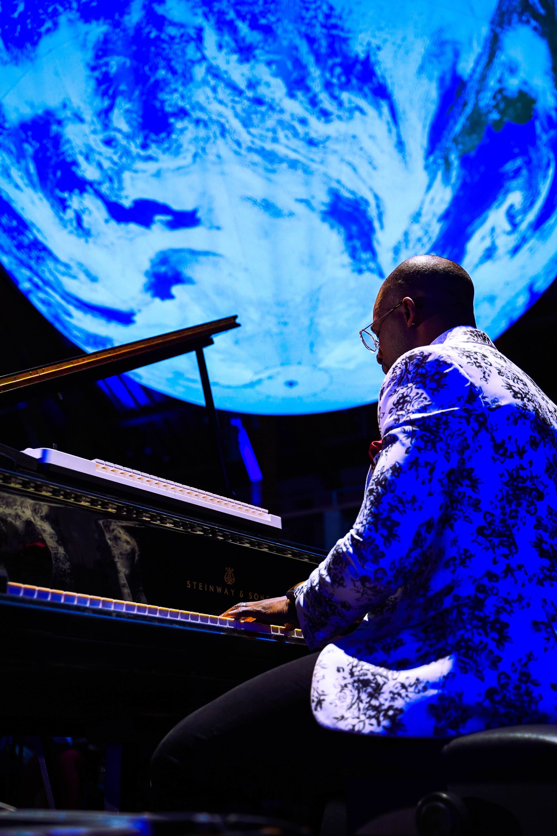 Pianist playing a grand piano, back to the camera. Earth-like blue background.
