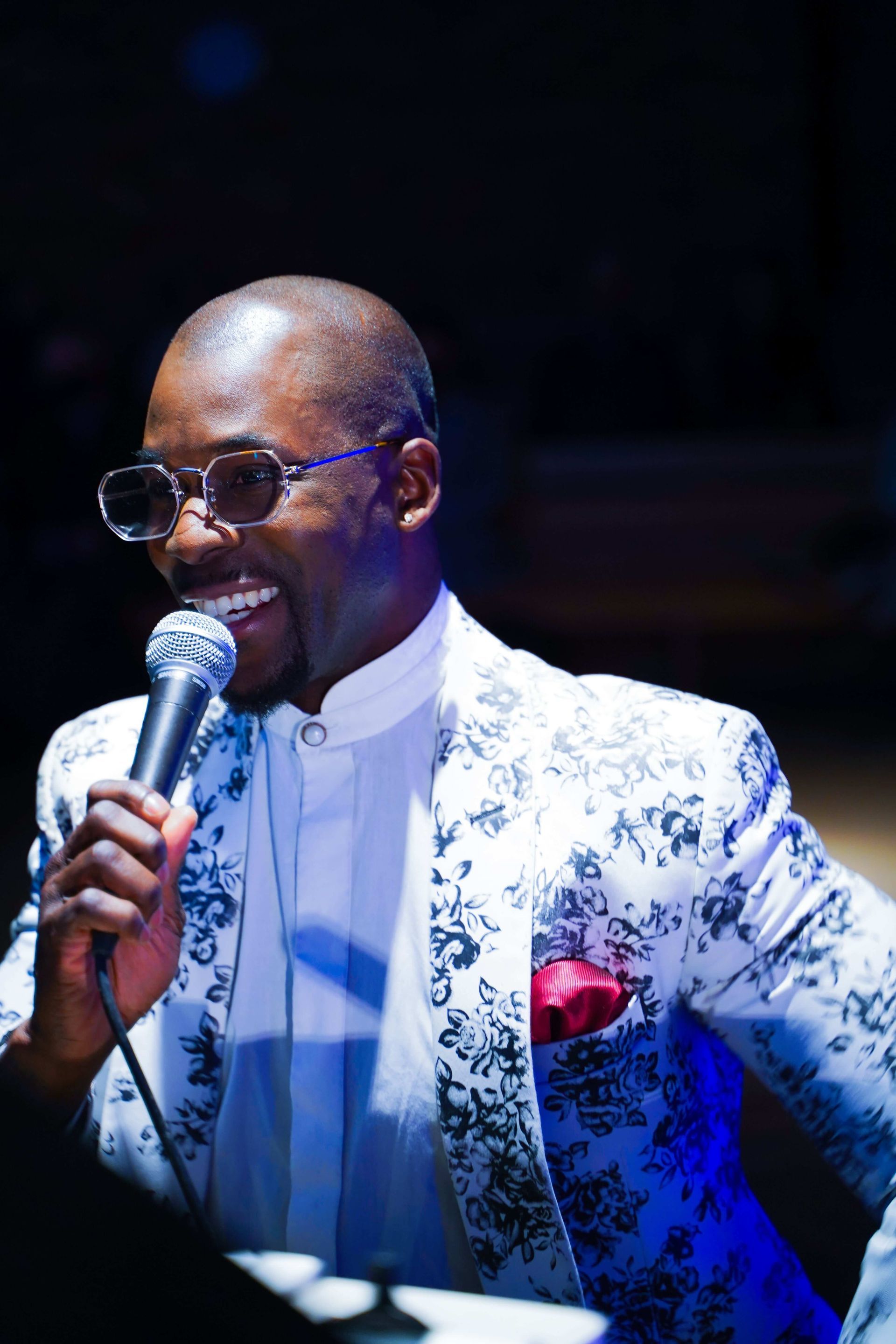 Man in floral jacket sings into a microphone. Smiling, wearing round glasses, indoors.