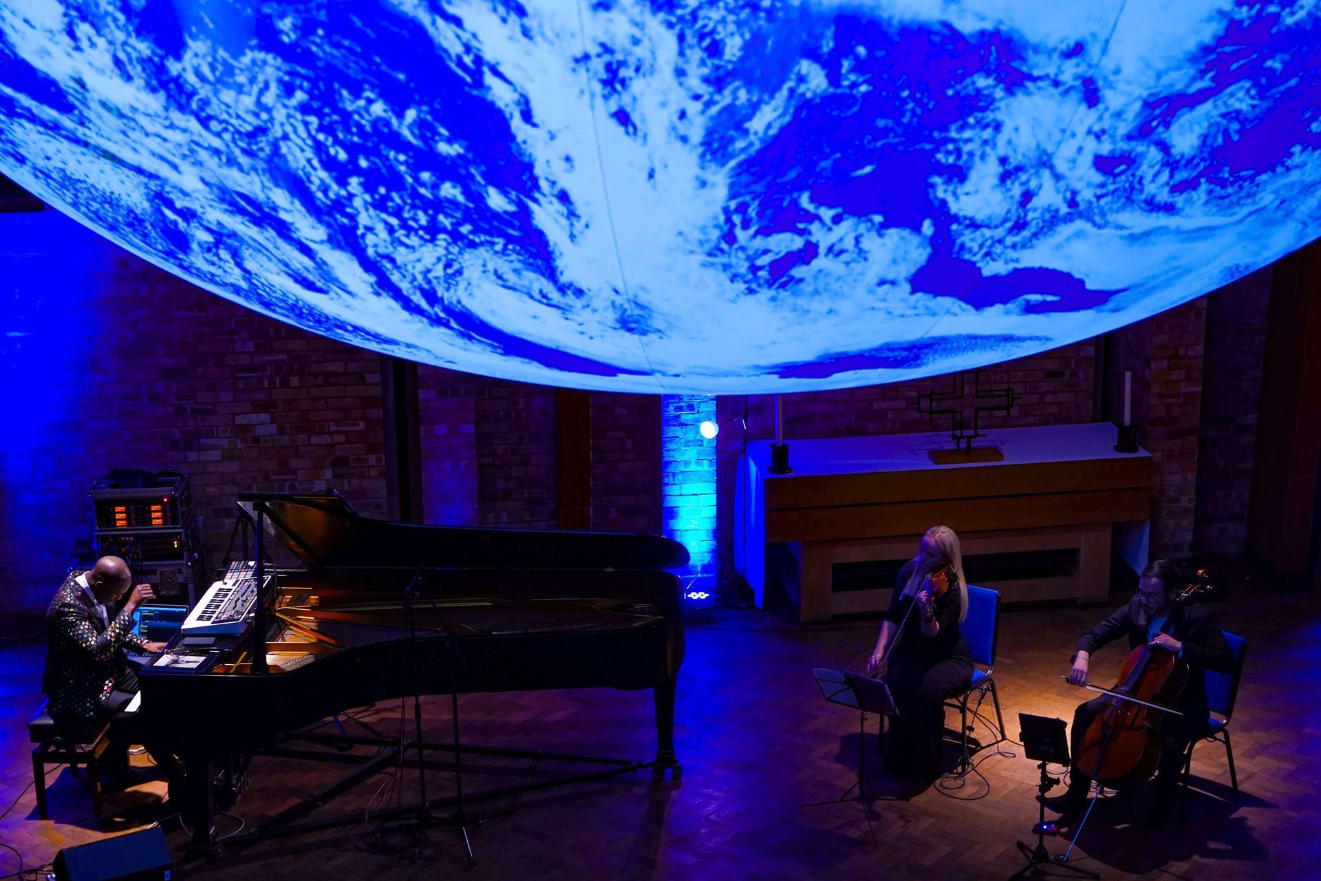 A piano and cello trio performs under a projected image of Earth. Blue lighting fills the space.