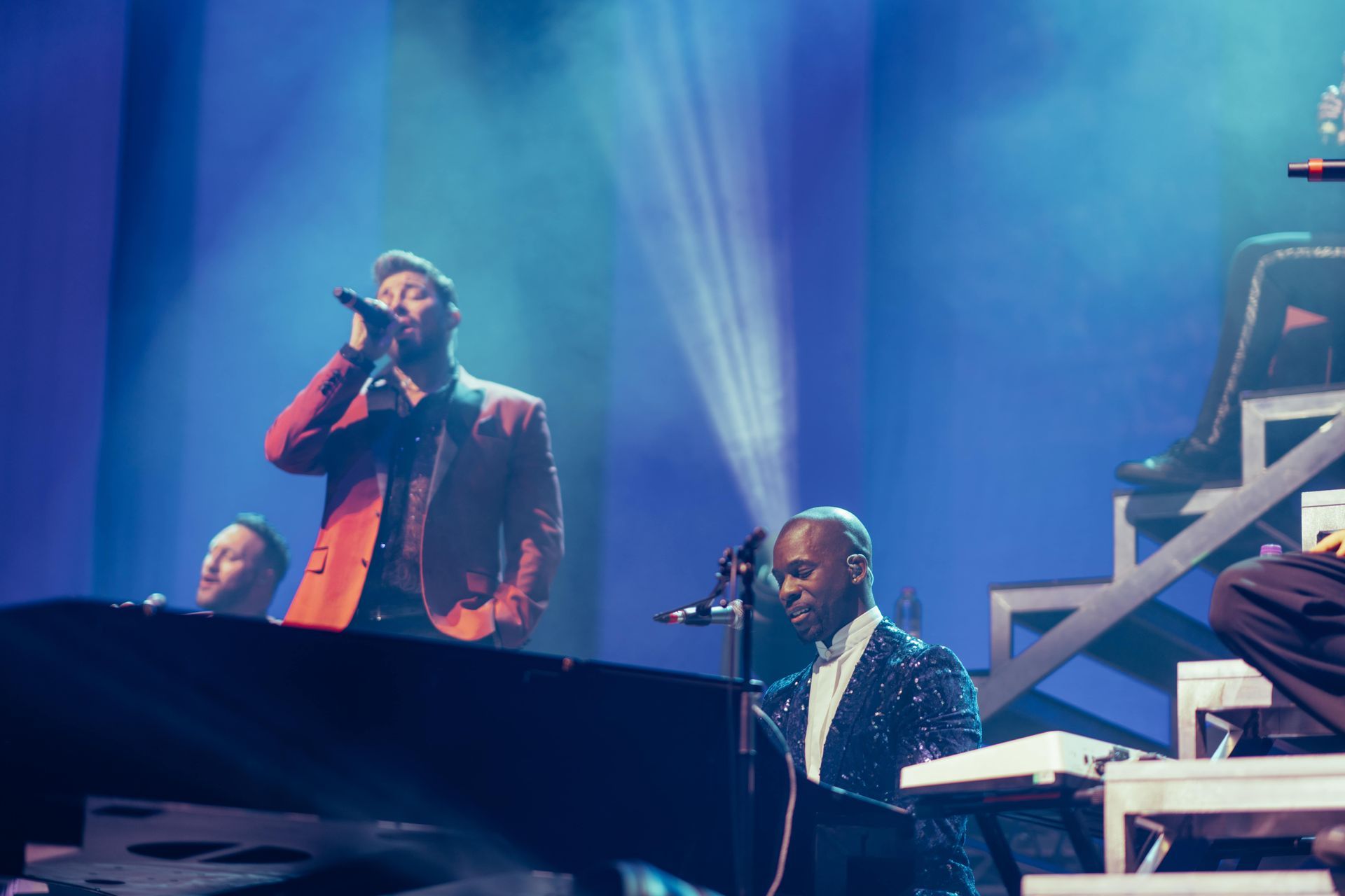 Band performing on stage, singer in red jacket, keyboardist in sparkly jacket, blue stage lights.