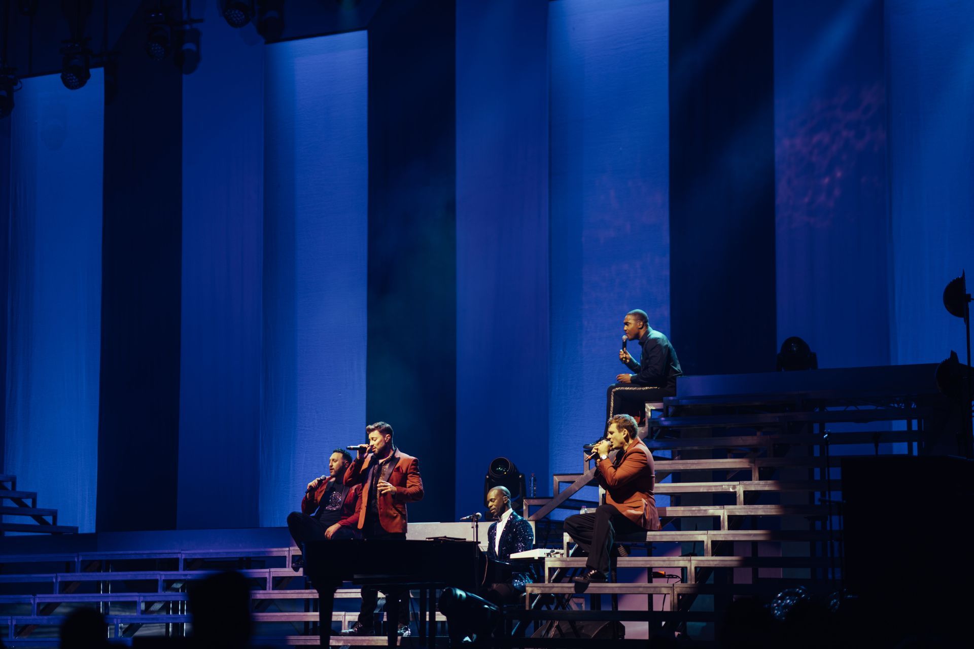 Four people performing on a stage with blue-lit background. Two singers stand, one sits, and one plays piano.