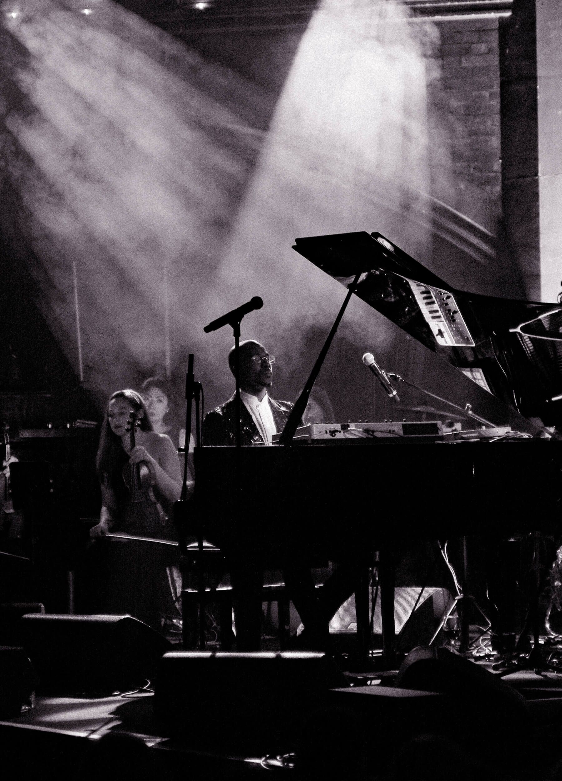 Piano player performing on stage with a vocalist, under a spotlight.
