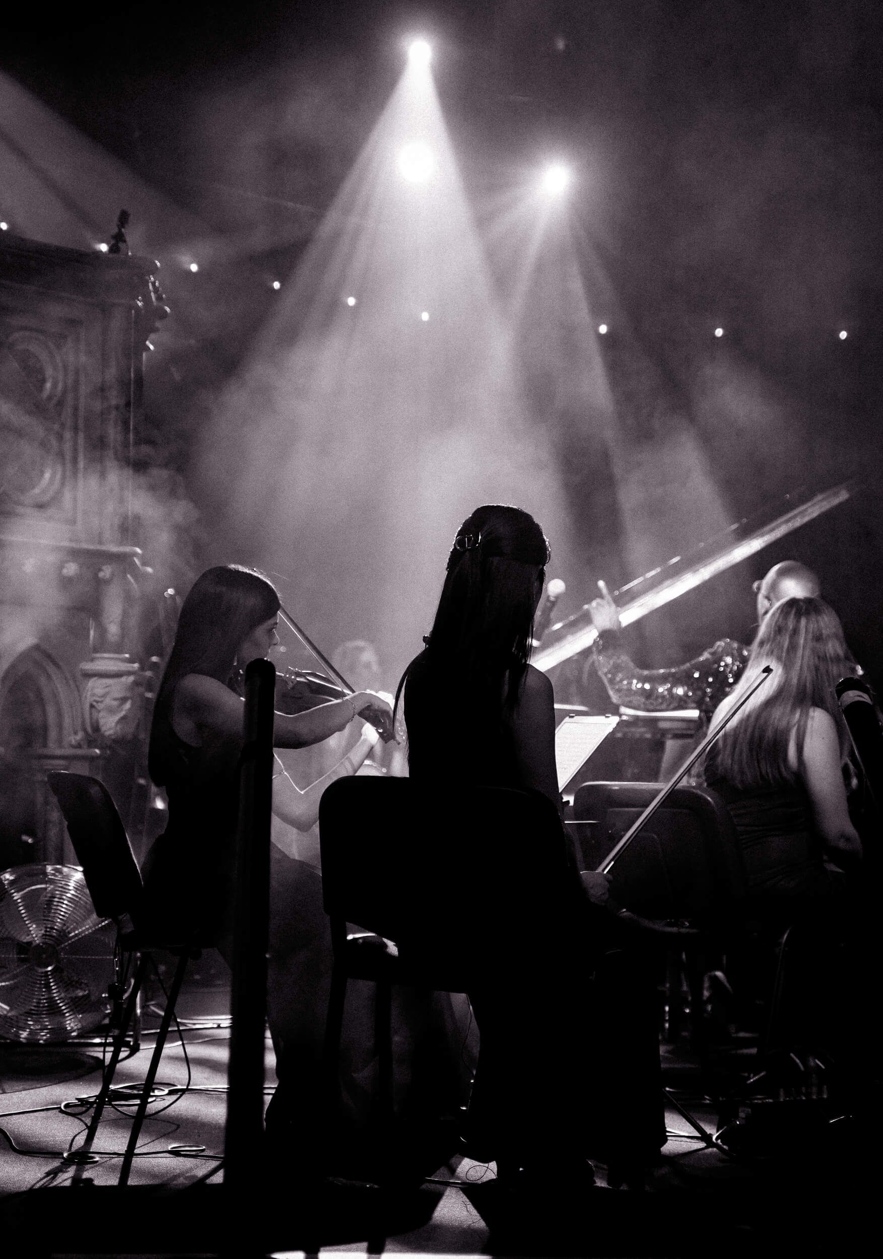 Concert scene: Silhouetted musicians on stage, illuminated by spotlights. A violinist and keyboard player are visible