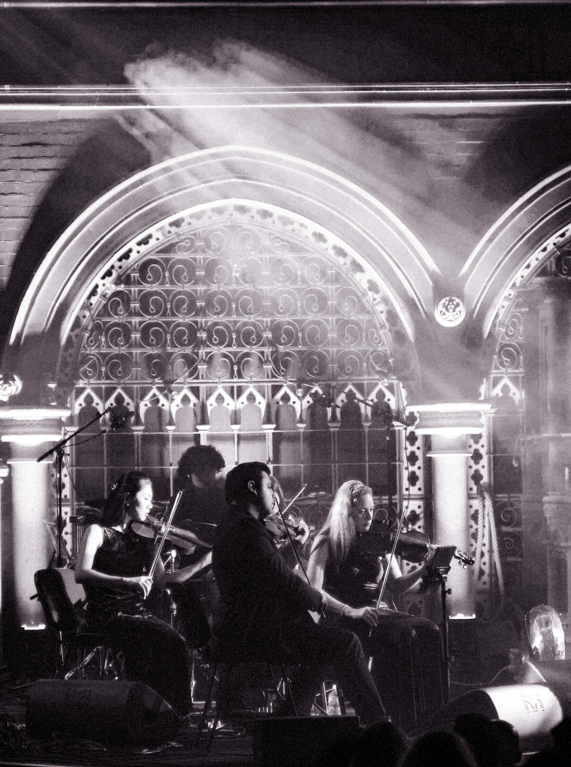Band performing on stage in front of arched architectural backdrop with stage lights.