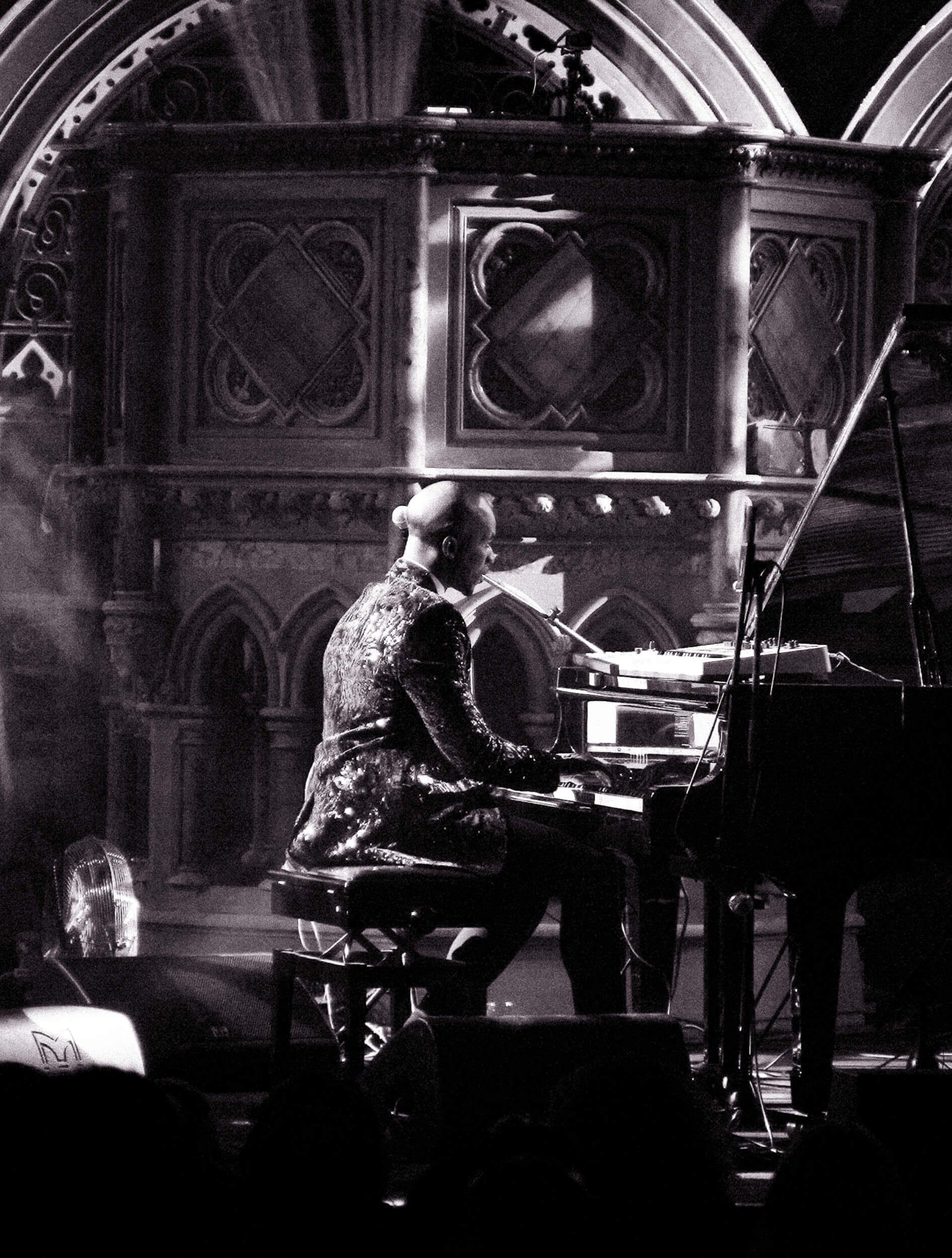 Pianist in a glittery suit plays a grand piano, back to the viewer, in front of a dark, gothic-style organ.