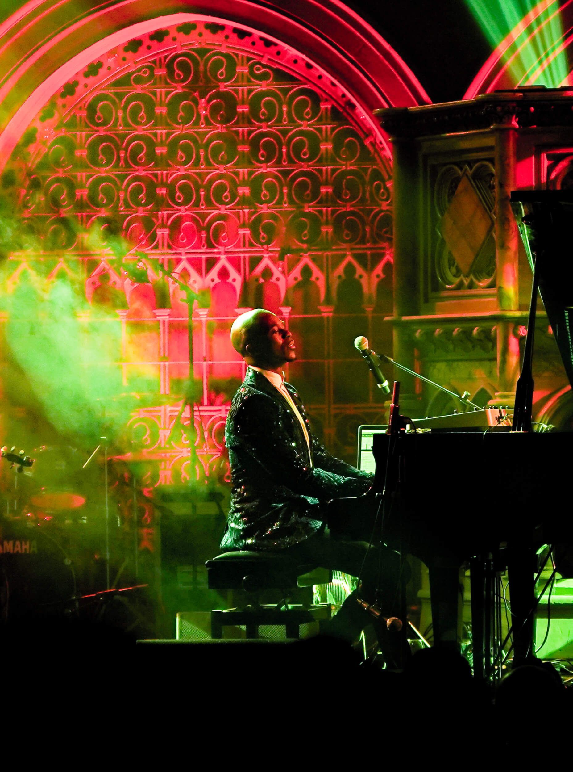 Man playing piano on stage, bathed in red and green light. Decorative background.