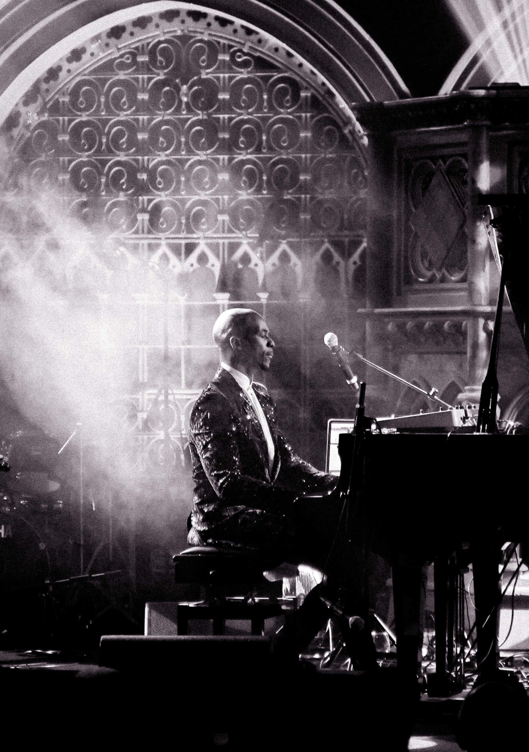 Man playing piano on stage, illuminated. Gothic backdrop. Black and white.