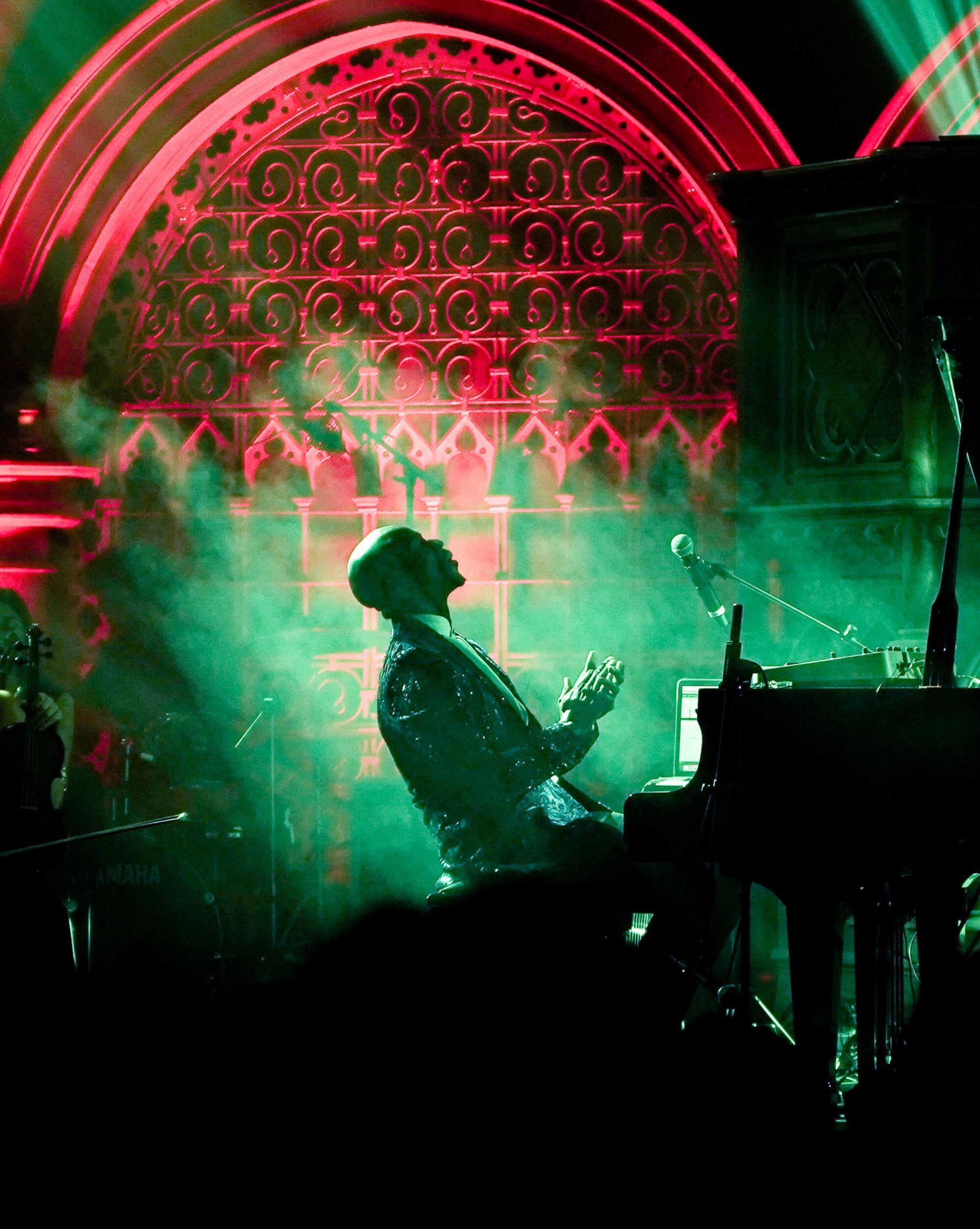 Pianist in a dark, stage-lit setting. Green and red lights illuminate a decorative window as the musician plays.