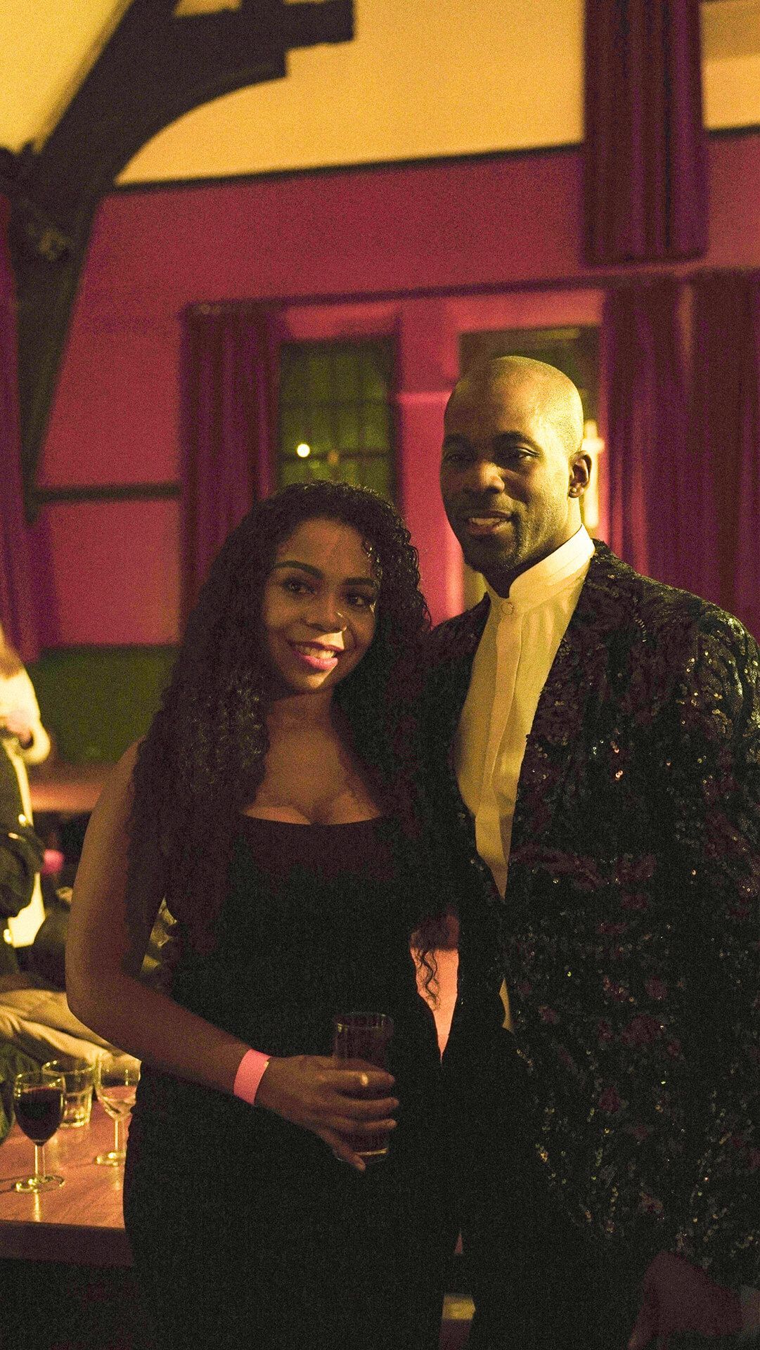 Couple smiling, posing together indoors; woman in black dress, man in sparkly jacket.