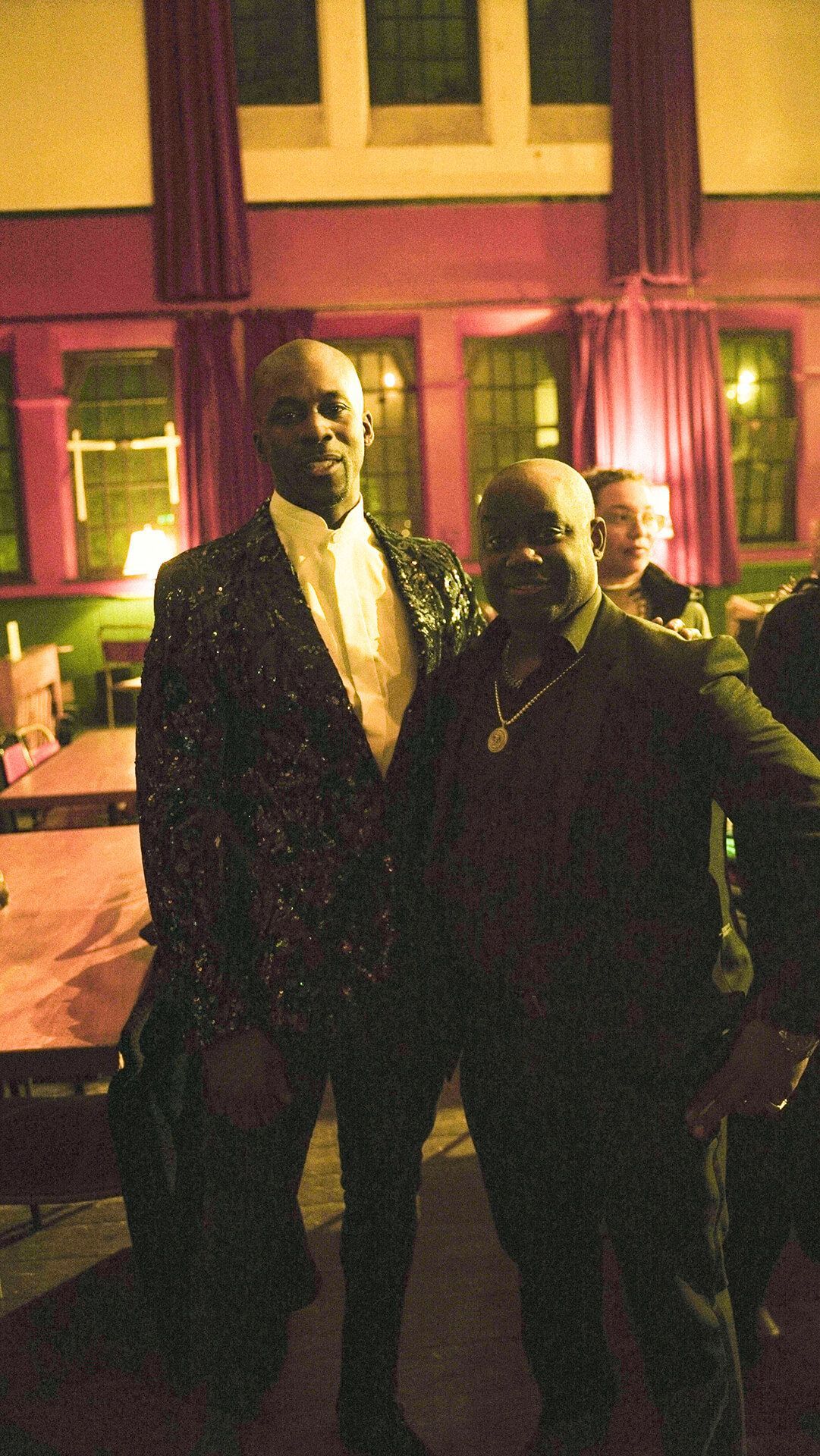 Two men pose in front of a red-curtained window, one in a sparkling suit, other in dark clothing.