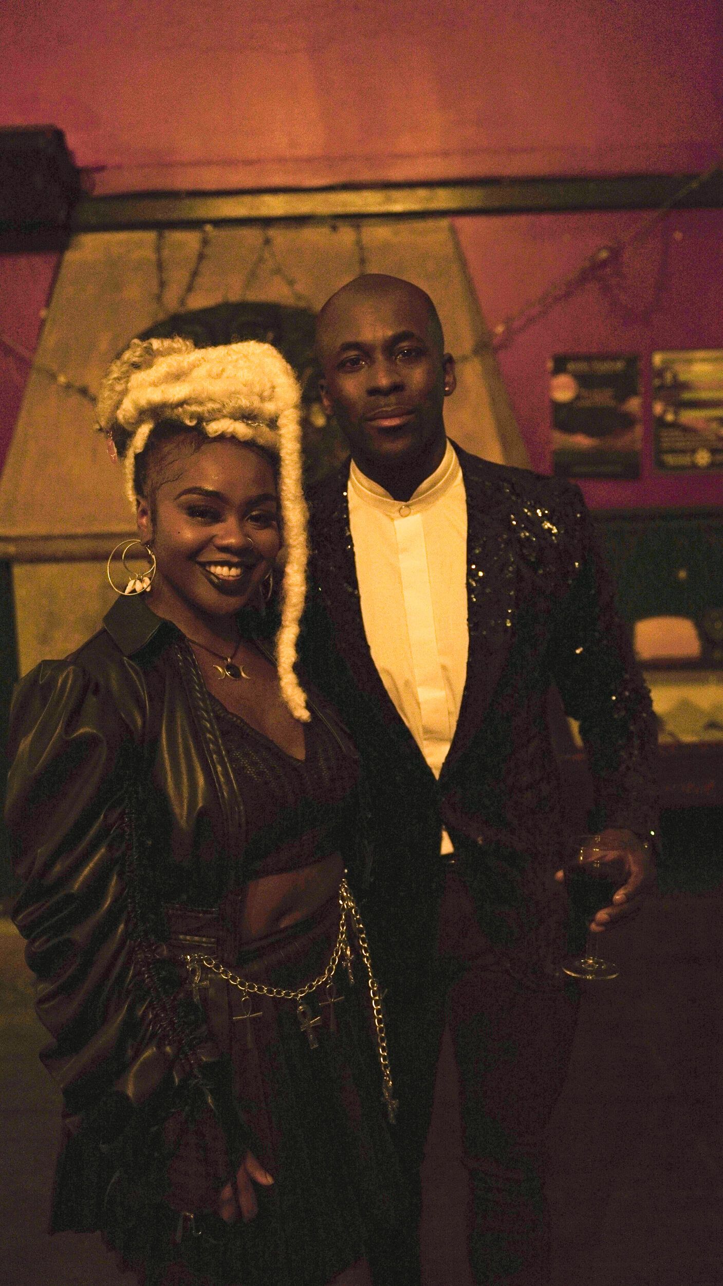 Couple posing indoors; woman with blonde dreadlocks, man in suit. Dark setting, smiling.