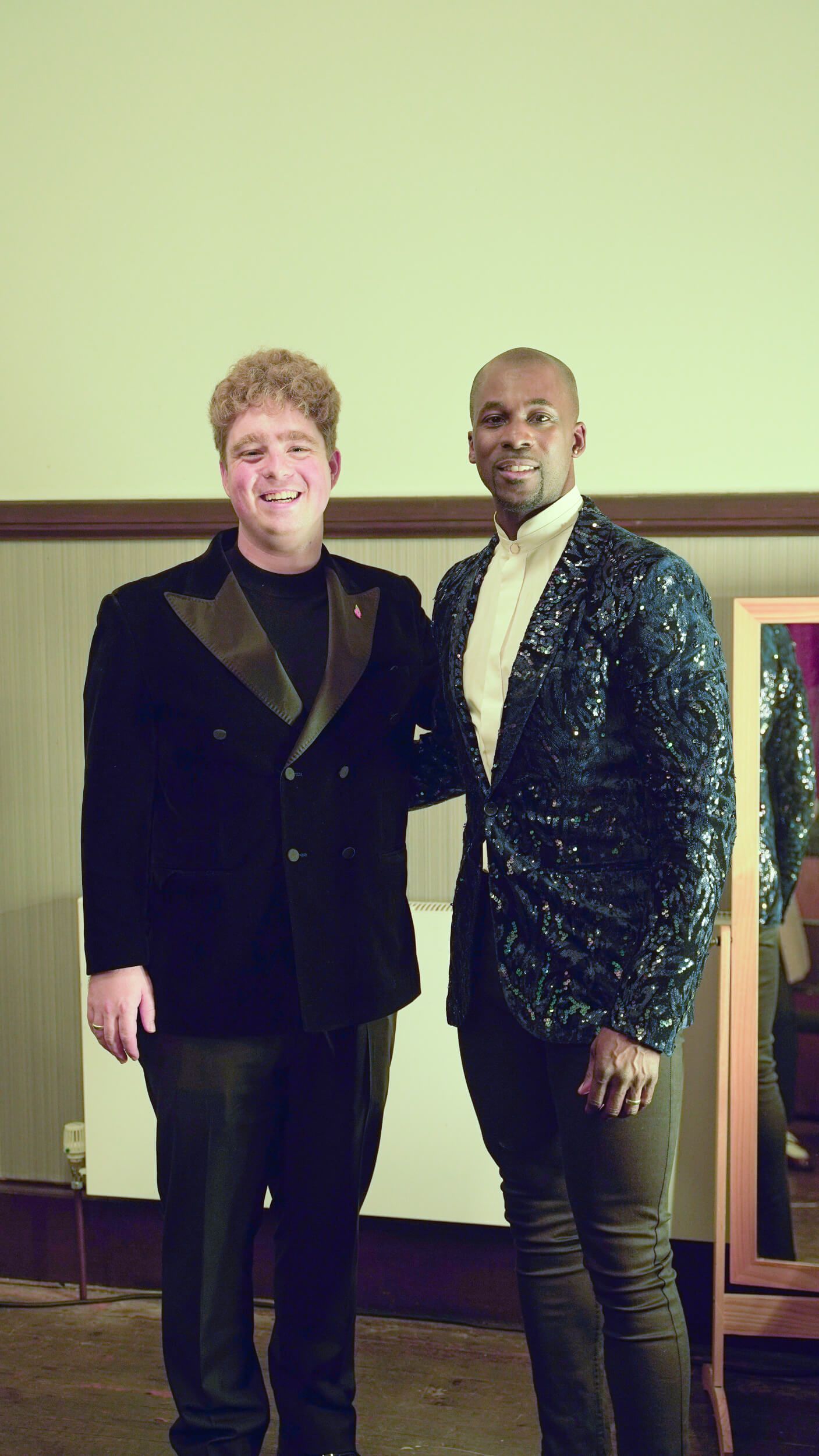 Two people in formalwear pose, one in a black blazer, the other in a sequined blue jacket. They stand indoors.