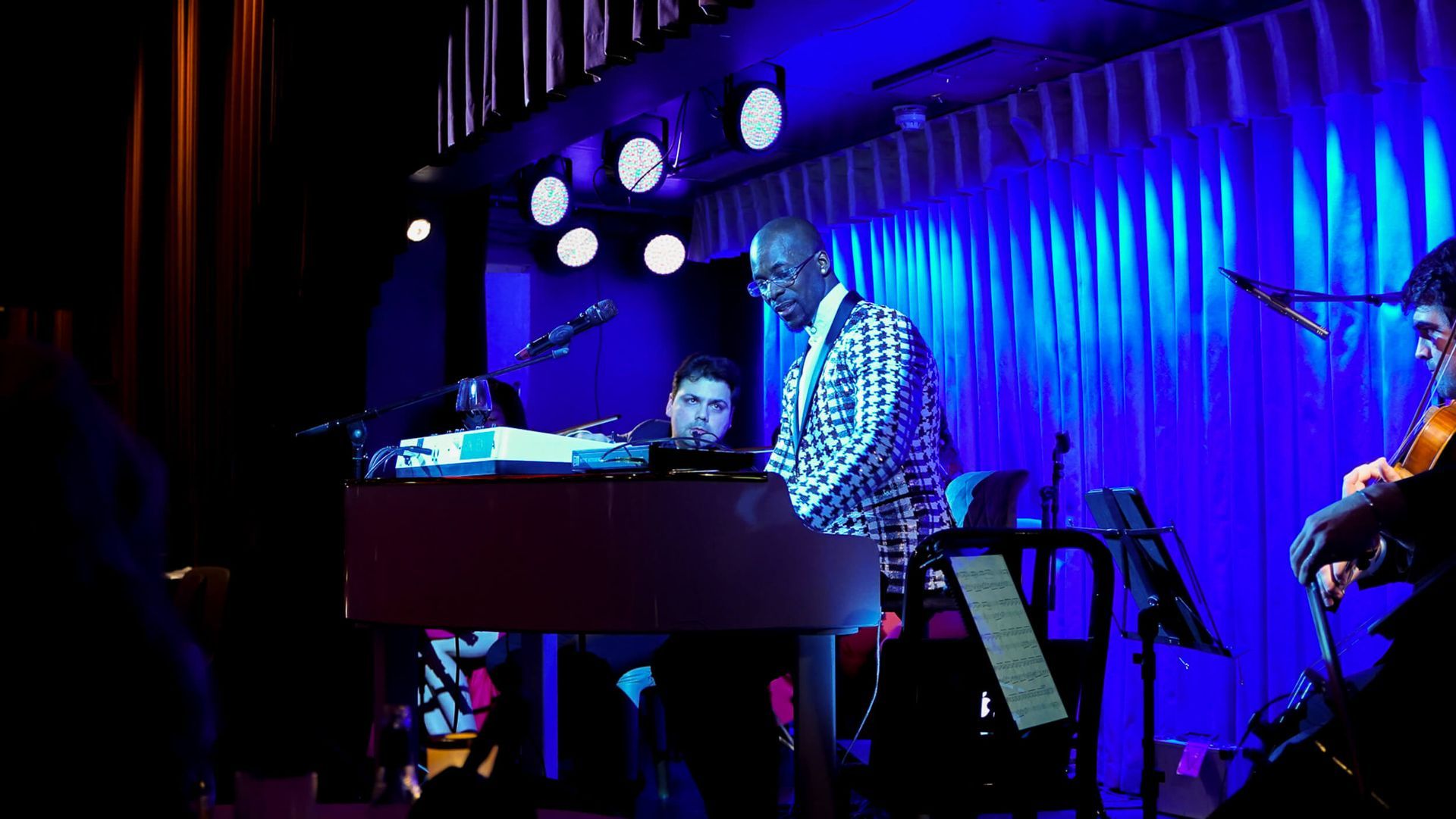 Keyboard player in patterned suit performs on stage with band, bathed in blue light.