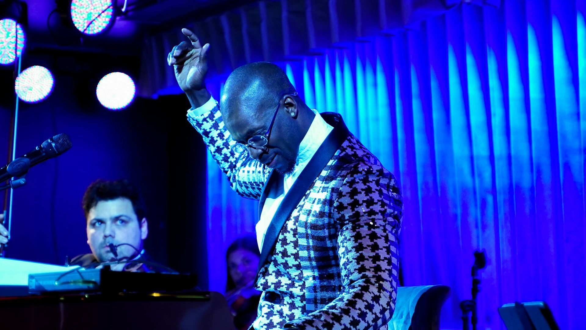 Man in sequined jacket singing, hand raised, on stage with keyboardist under blue lights.