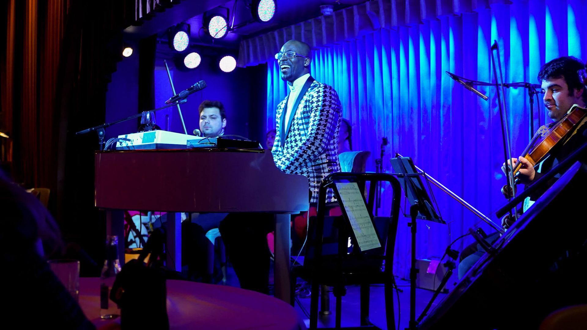 Band performs on stage under blue lighting; singer in checkered jacket, keyboardist, violinist.