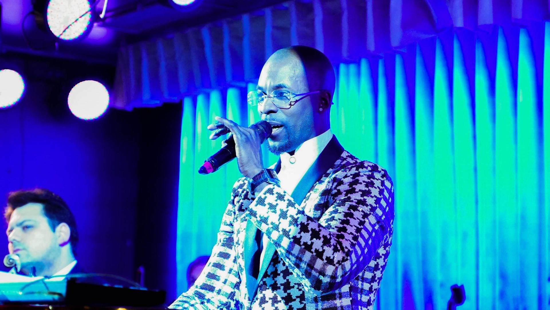 Man singing into microphone on stage, wearing a black and white patterned jacket. Blue lighting.