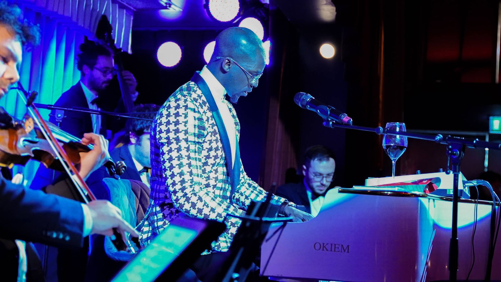 A musician in a patterned jacket plays a piano on stage with a band, lit by stage lights.