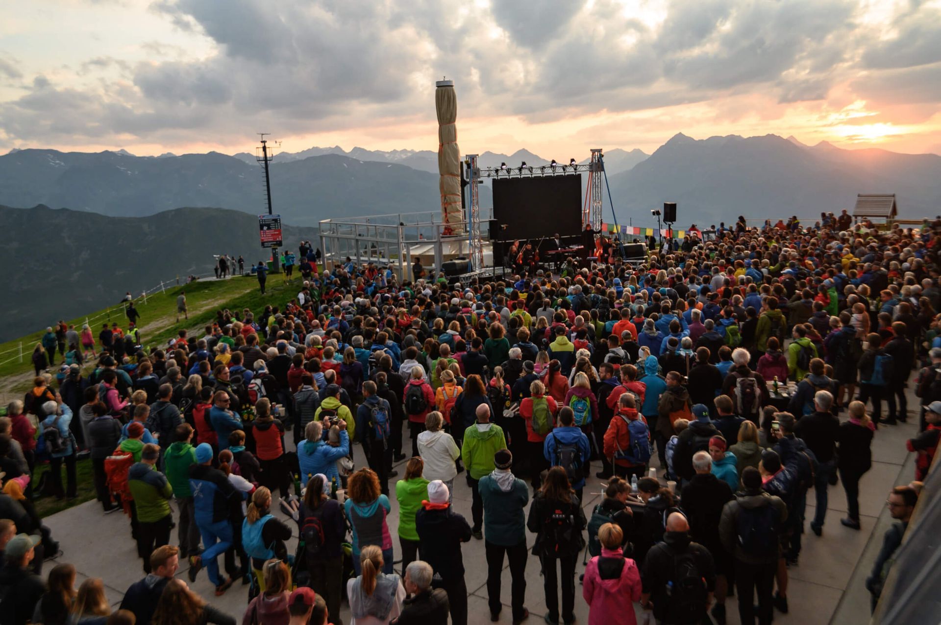 Large crowd gathered outdoors at a mountain summit; stage set up with backdrop; sunset behind mountains.