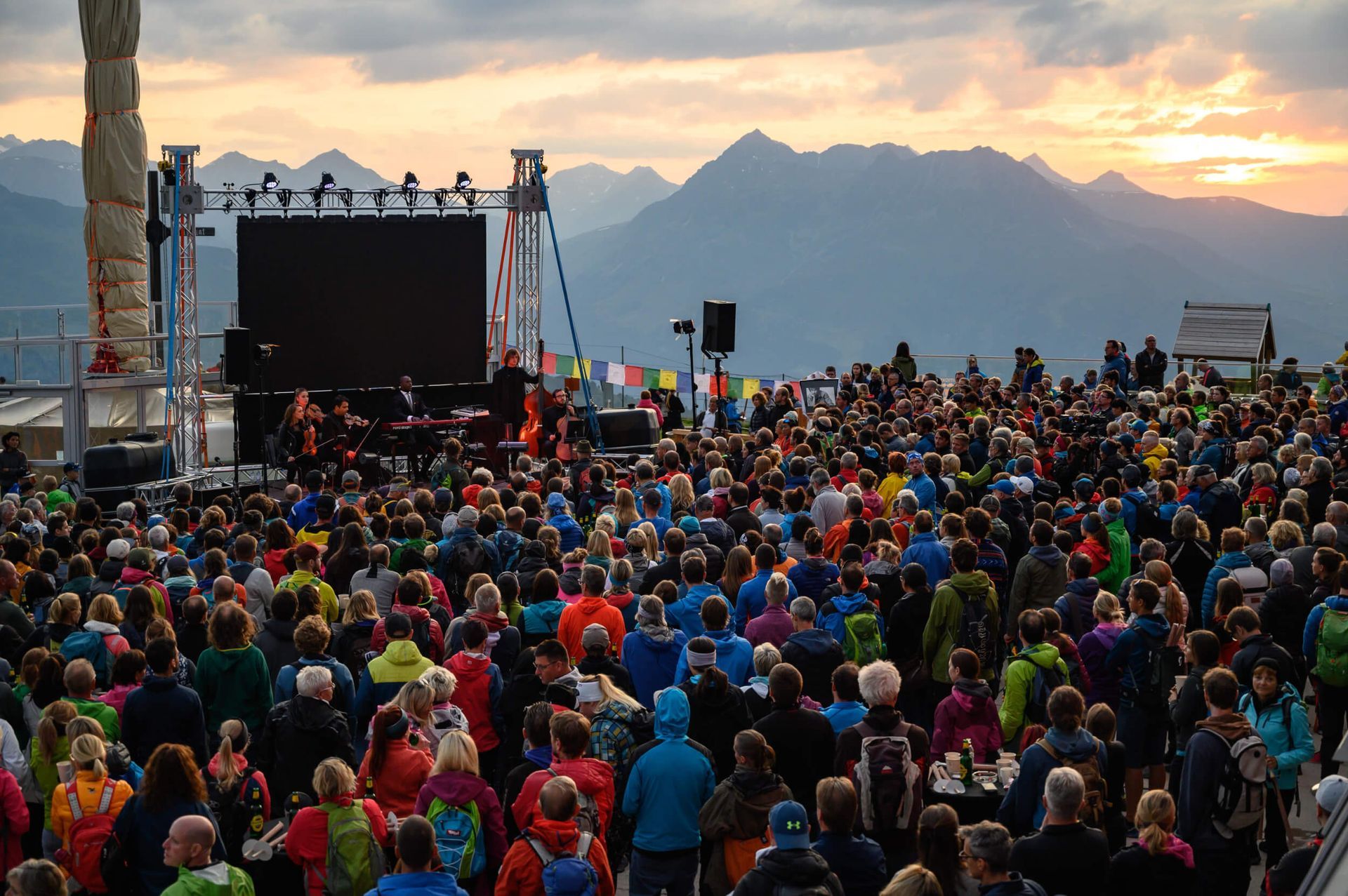 Large crowd at outdoor concert with stage, mountains, and sunset.