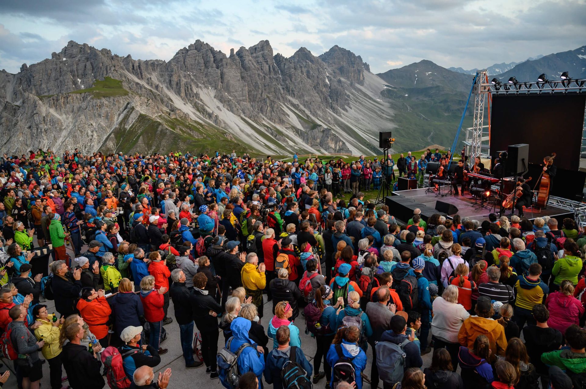 Large crowd at an outdoor concert on a mountain with a stage and mountains in the background.