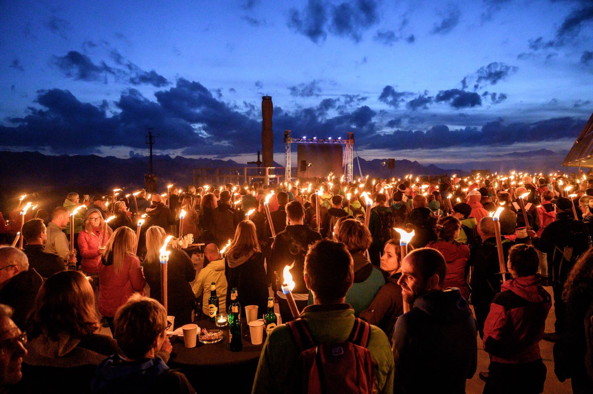 Crowd of people holding torches at dusk; event outdoors with a stage and dark sky.