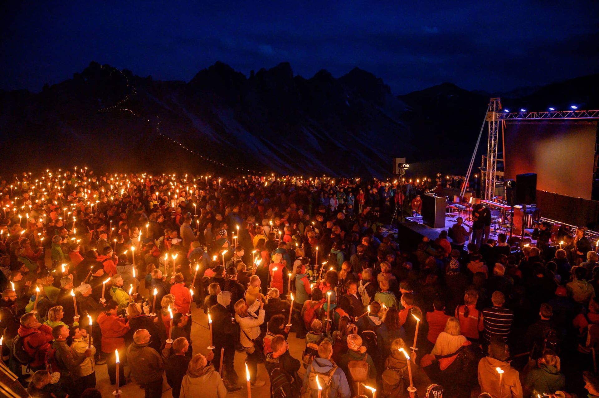 Large crowd holding torches at night, gathered near a stage in front of a mountainous backdrop.