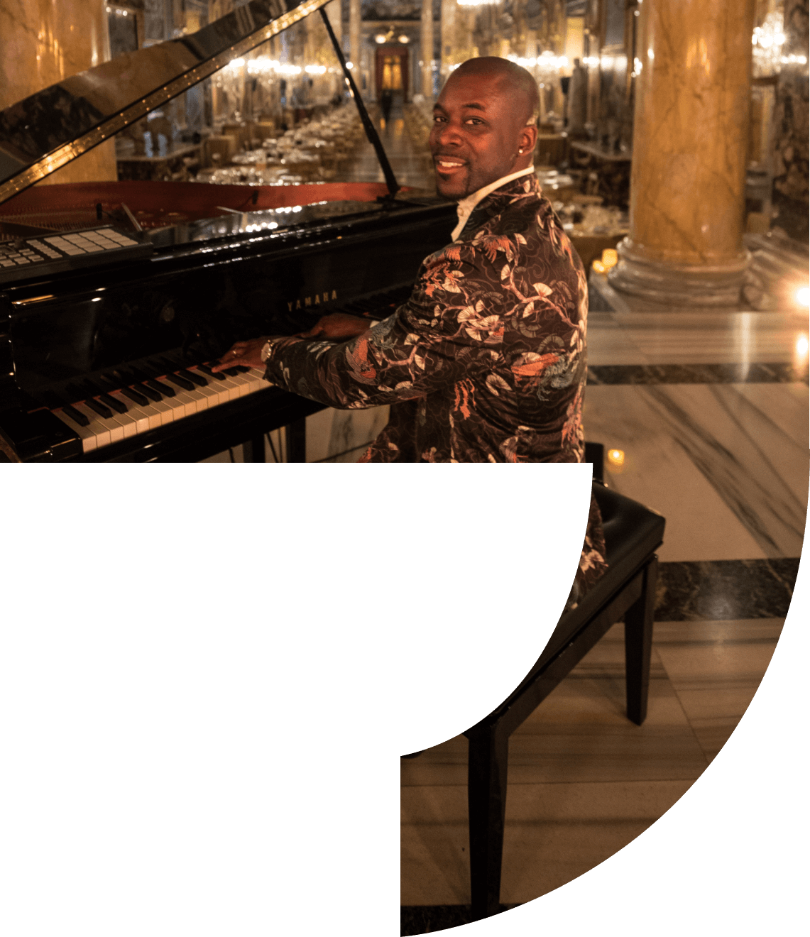 Man playing a grand piano in an ornate room, smiling, wearing floral blazer.